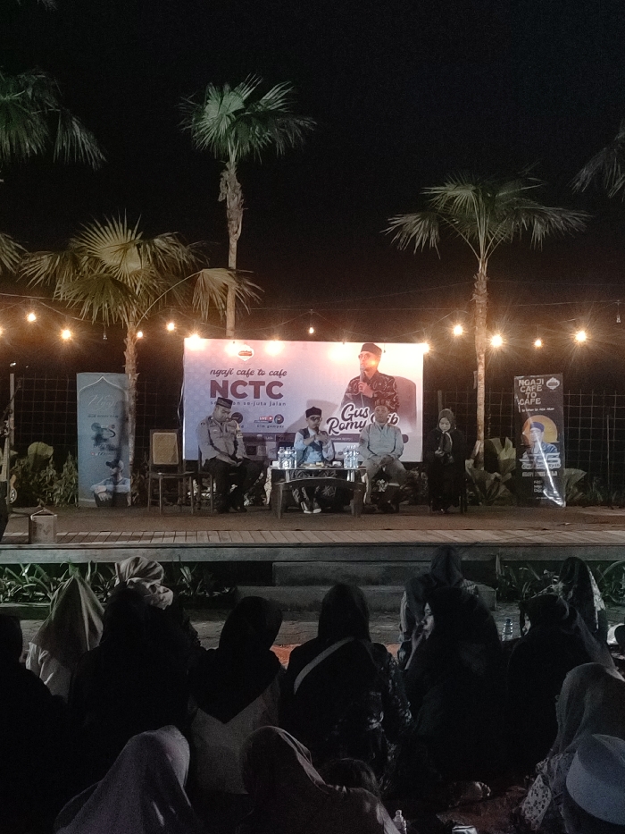 The image shows a group of people seated in front of a stage in an outdoor setting in the city of Purwosari, Indonesia, at coordinates -7.747, 112.737. The stage is decorated with a large banner that reads "ngaji cafe to cafe NCTC" in yellow, white, and red lettering. The banner also has a picture of four people with microphones, and a small banner with the words "Gus Romy" in white. There are three palm trees on the left and right sides of the stage. The stage is illuminated by strings of light bulbs hanging from the trees above. The people in the audience are wearing black and gray clothes, with their faces obscured. The environment is dark, with dim lighting from the strings of lights. There is a small wooden table on the stage, on which are placed several water bottles, microphones, and other items. The people on the stage are speaking into microphones. The image appears to be taken at night.
