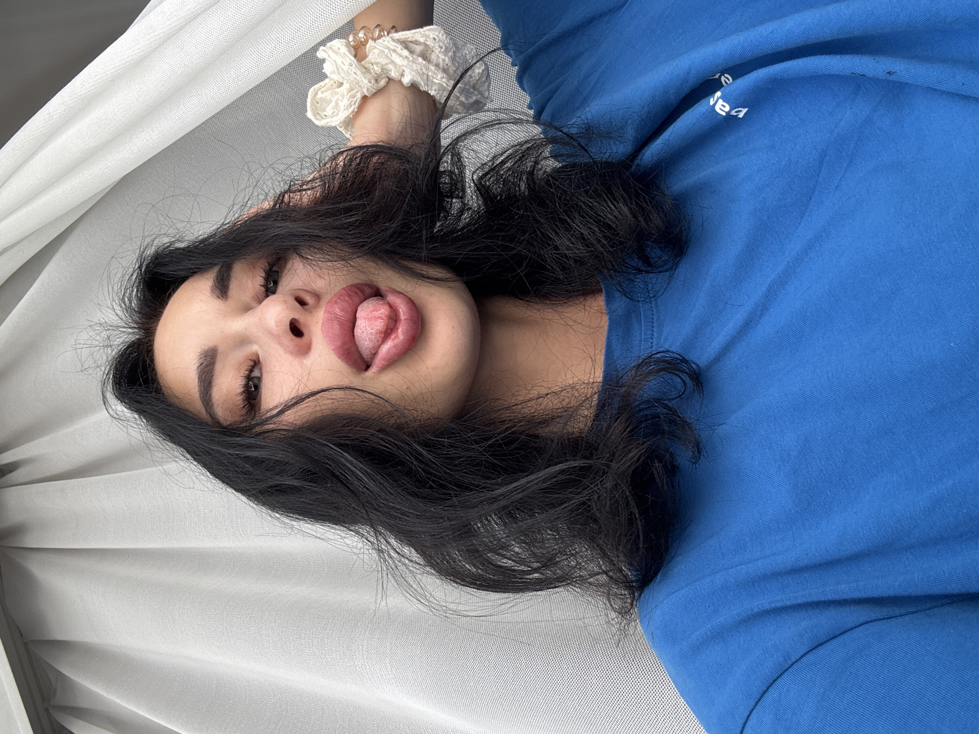 A close-up selfie of a woman with black hair and full lips sticking her tongue out. She is wearing a blue t-shirt and has a white scrunchie on her left wrist. The background consists of a white, sheer curtain. There is no indication of the city of Chaikovsky, Russia in the image.
