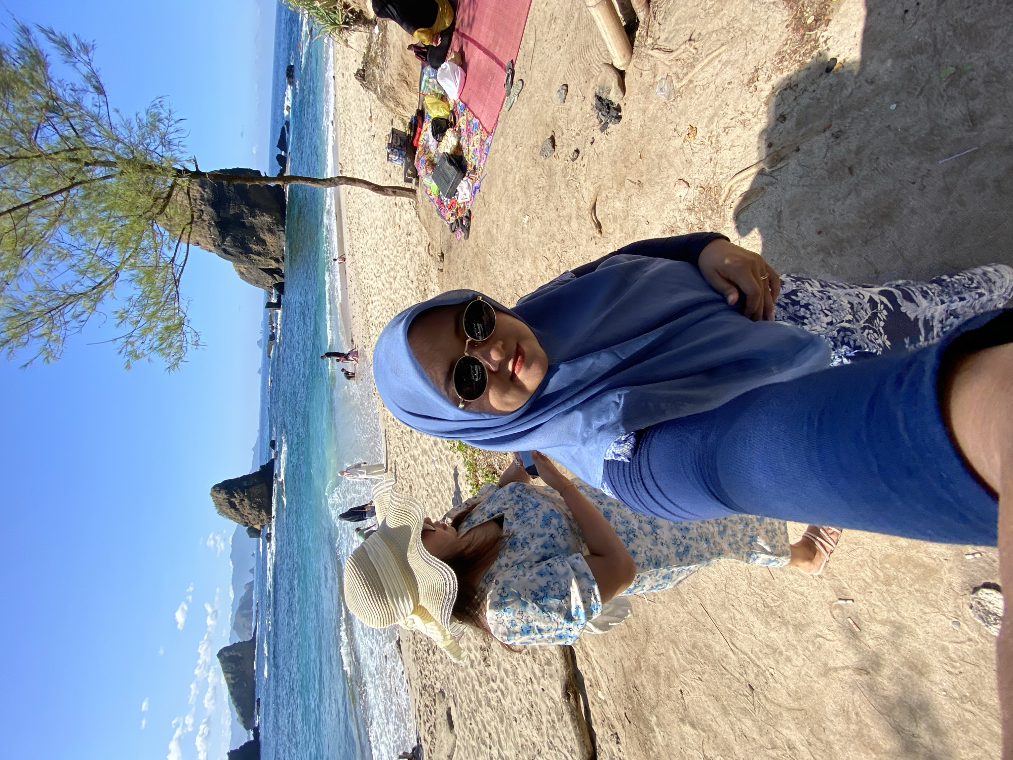 Two women are visible in the image. One woman wears a blue hijab and sunglasses, while the other wears a white sun hat and a light blue floral dress. The woman in the hijab appears to be holding a cell phone, possibly taking a selfie. They are located on a sandy beach near the ocean. In the background, there are rock formations, a few other people on the beach, and some personal belongings such as a blanket and bags. The picture was taken in Unknown, Indonesia.