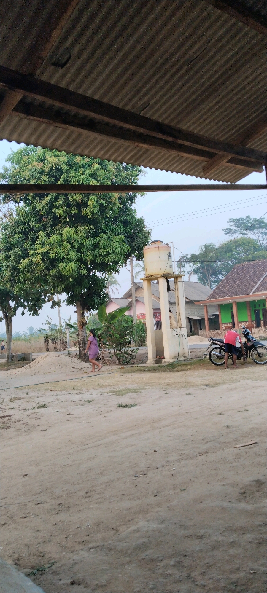 This image depicts a dusty outdoor scene in Tanjung Ratu Ilir, Indonesia. In the foreground, a weathered corrugated metal roof overhangs the view. To the left, a large, leafy green tree stands prominently. A woman in a purple dress is walking on the dirt path in front of the tree. In the center of the image, a tall, white water tank on stilts is visible, with pipes running down its side. To the right of the water tank, a man in a red shirt and dark shorts is standing next to a black motorcycle, appearing to adjust something on it. Behind these elements, a residential building with a dark roof and a green section is situated. The ground is predominantly composed of dirt and sand, with some scattered debris. The sky is overcast, suggesting a hazy or early morning/late afternoon light. The overall impression is of a rural or semi-rural Indonesian village setting.