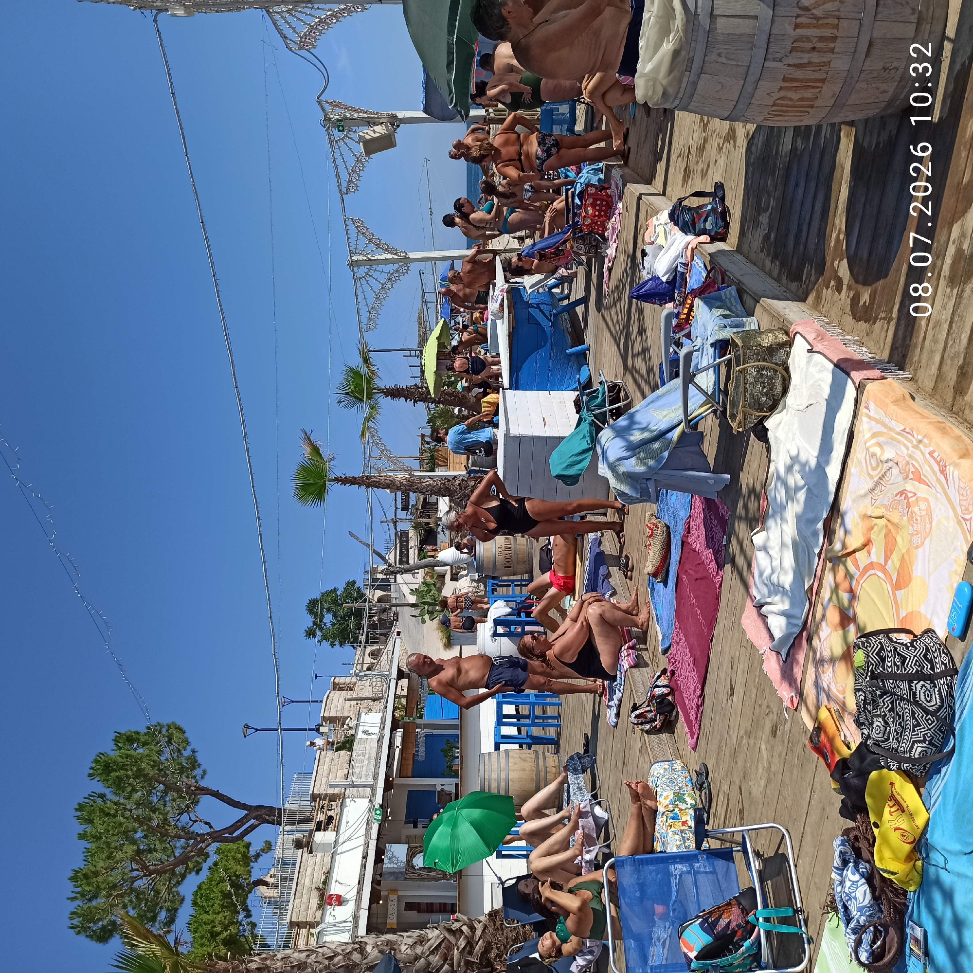 The image depicts a crowded beach scene in Trani, Italy, on a sunny day. People are seen relaxing on lounge chairs and towels laid out on a wooden boardwalk. Several individuals are sunbathing, some are standing and chatting, and others are in the process of eating or drinking. There are also some umbrellas providing shade. In the background, a building with a sign that says "CASSA" is visible, along with a bar and some palm trees. The overall atmosphere is one of leisure and recreation.