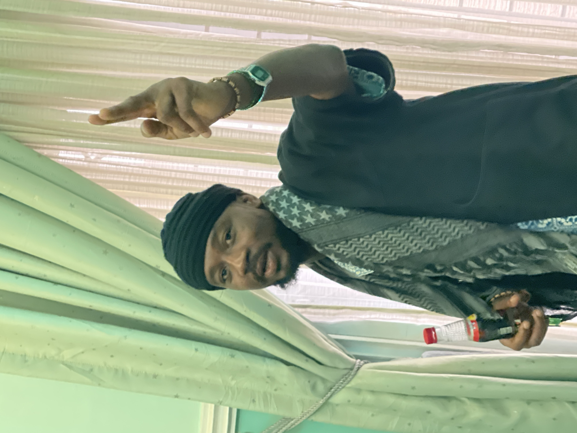 A man with dark skin, a black beanie, and a gray and white patterned scarf is pointing his left hand. He is wearing a watch and a bracelet. He is holding a small water bottle in his right hand. He is standing in front of a window with light-colored curtains. The location is Gura Top, Nigeria.