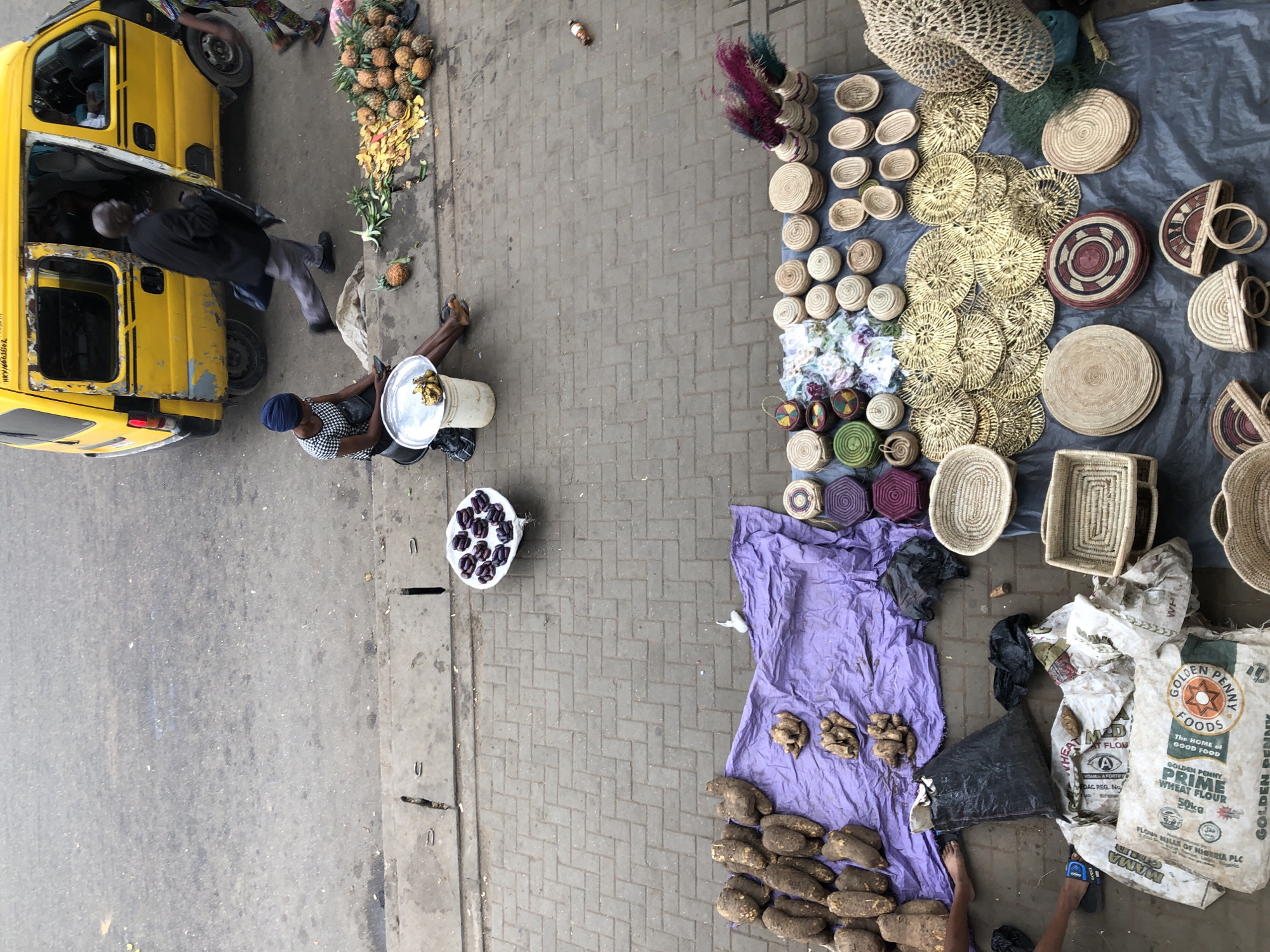 An aerial shot shows a street market scene in an urban area. A yellow van with an open door is parked on the asphalt road. A man in a black jacket and gray pants exits the van, carrying a dark bag. A woman with a blue headscarf and a checkered shirt sits on a concrete step near the van, looking at her phone. A pile of pineapples and banana peels is on the ground next to her. On the paved sidewalk, a purple cloth is spread out with a collection of large, brown root vegetables. Further down the sidewalk, a variety of woven straw baskets and bowls are displayed on a dark cloth. Several people's legs and feet are visible at the bottom of the frame, suggesting they are shopping or working at the market. The environment appears to be a busy marketplace, with goods being sold and people engaged in transactions.