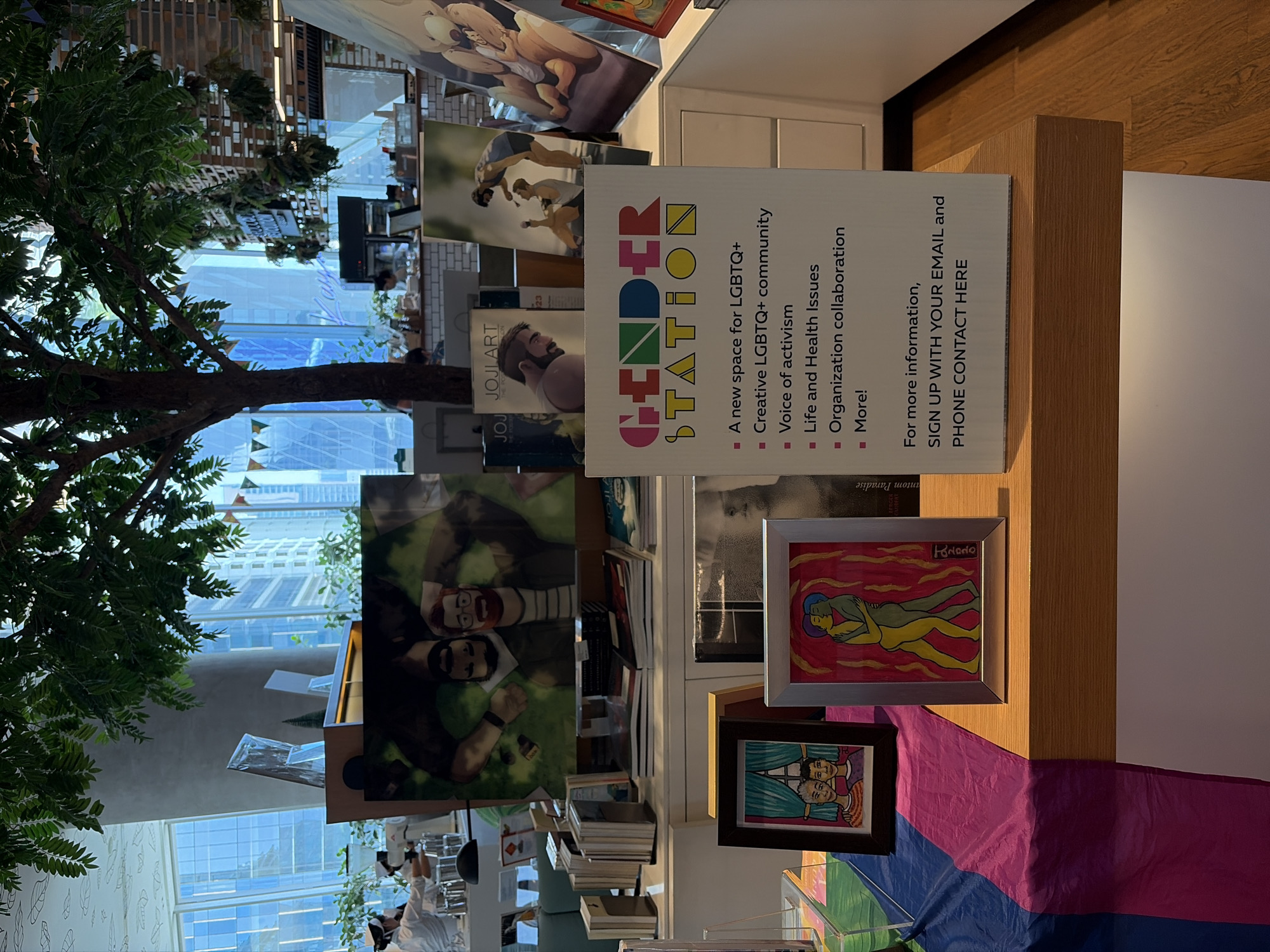 The image shows an interior space, possibly a bookstore or community center in Bangkok, Thailand, related to the LGBTQ+ community. The area is decorated with various images and artwork, including framed art and a large poster for "Gender Station," highlighting its mission as a creative space for LGBTQ+ individuals and activism. A bi flag is draped below the artwork. Books are stacked on shelves, and part of a tree is visible in the foreground, implying a connection to nature or an outdoor view. The setting appears to be in a modern building with large windows, allowing natural light to illuminate the space.