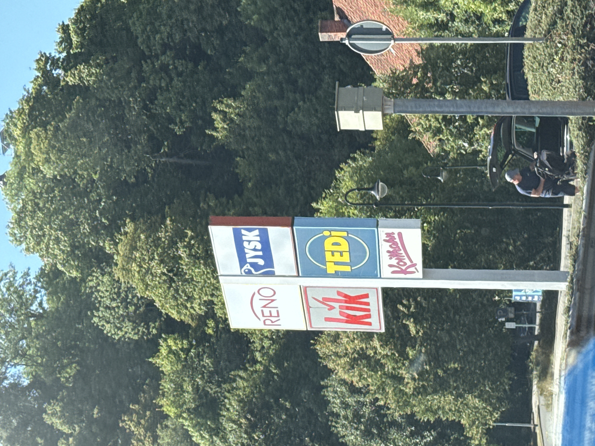 The image captures a street-level view in Herzberg, Germany, featuring a prominent advertising pylon displaying multiple store logos. The pylon shows four stacked signs: "RENO" in red lettering on a white background, "JYSK" with a bird graphic in blue and white, "kik" in red with a checkmark graphic, and "TEDI" in yellow within a blue circle on a blue background. Below the "TEDI" sign, a partially visible sign reads "Koithahn" in pink script on white.
To the right of the pylon, a speed camera is mounted on a pole. Further to the right, a red-tiled roof of a building is visible, along with a street sign. In the background, a dense forest of green trees covers a hillside under a clear blue sky.
In the foreground, a man wearing a blue t-shirt and a cap is seen next to the open trunk of a dark-colored car, appearing to interact with a stroller or baby carrier. A portion of a road and some bushes are also visible in the lower part of the frame. The scene suggests a commercial area situated near a natural landscape.