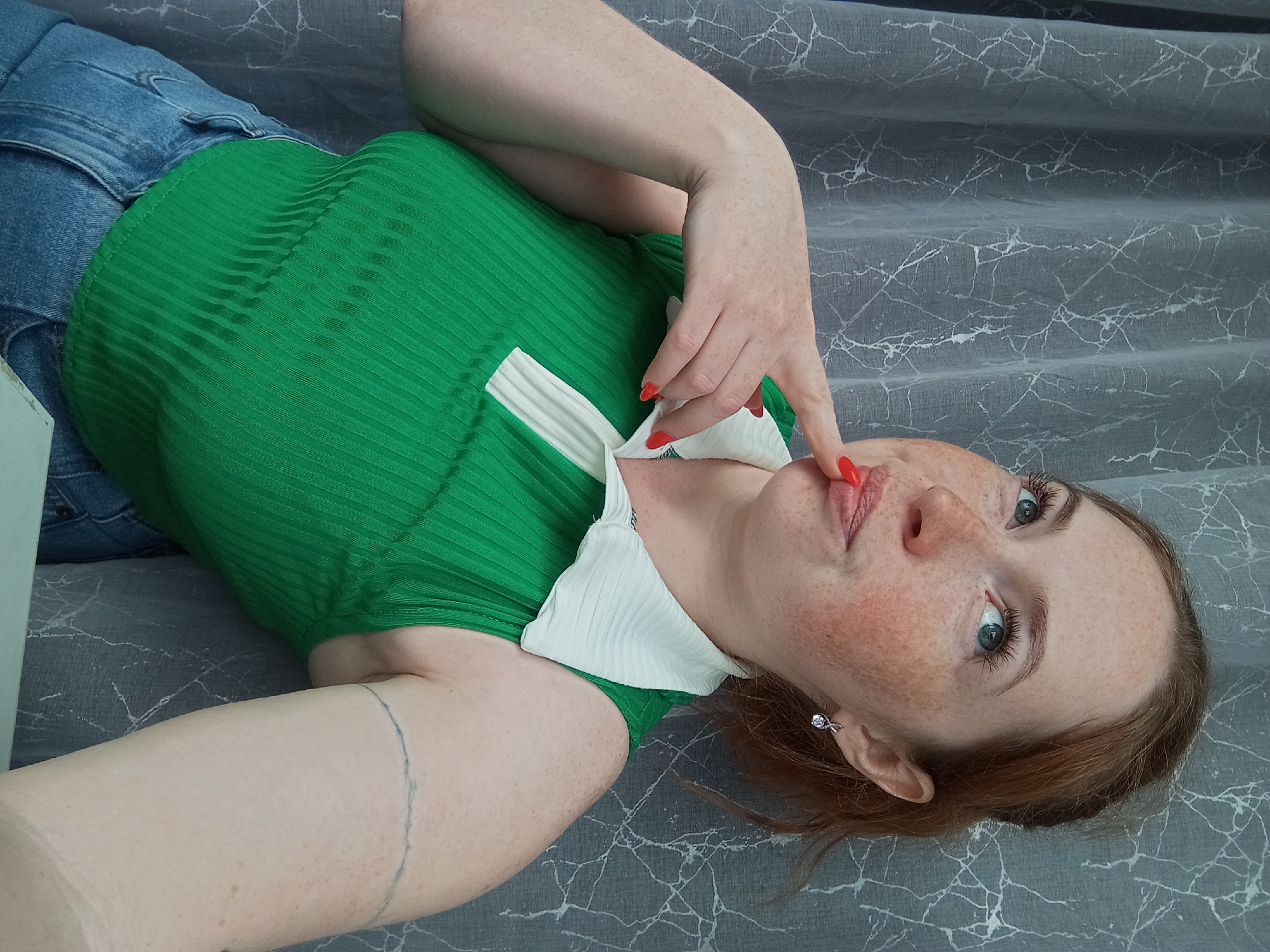 A freckled woman with red hair and blue eyes lies on a grey patterned surface, possibly a bed or couch, in Yurgamysh, Russia. She wears a green, ribbed, short-sleeved top with a white collar and blue jeans. Her fingernails are painted red. Her right hand touches her lips. The background is out of focus.