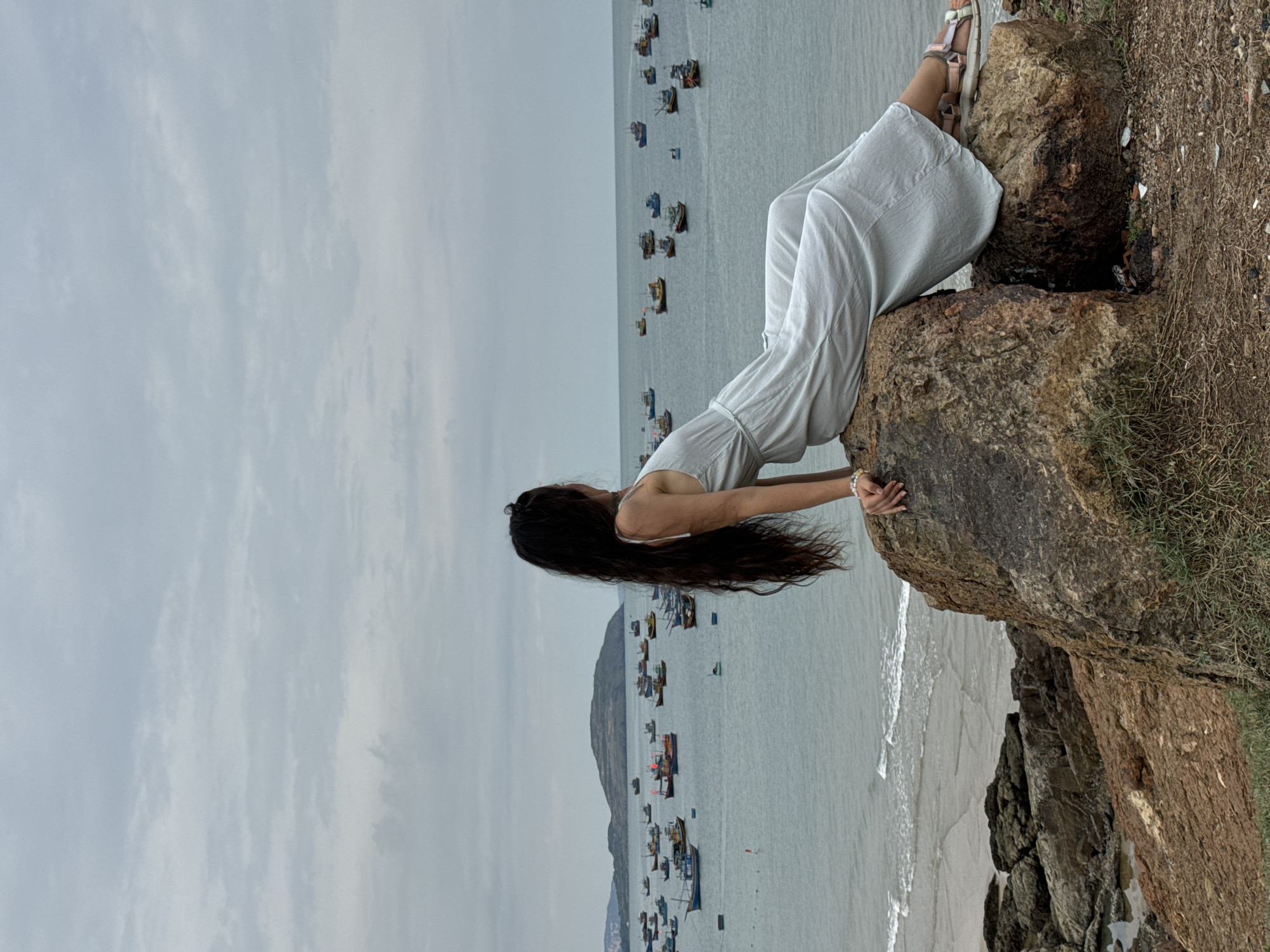 A woman in a light-colored dress sits on a large rock overlooking the ocean in Mui Ne, Vietnam. Her long, dark hair hangs down her back. Many small fishing boats dot the water. The sky is overcast. The rock is surrounded by sparse vegetation.