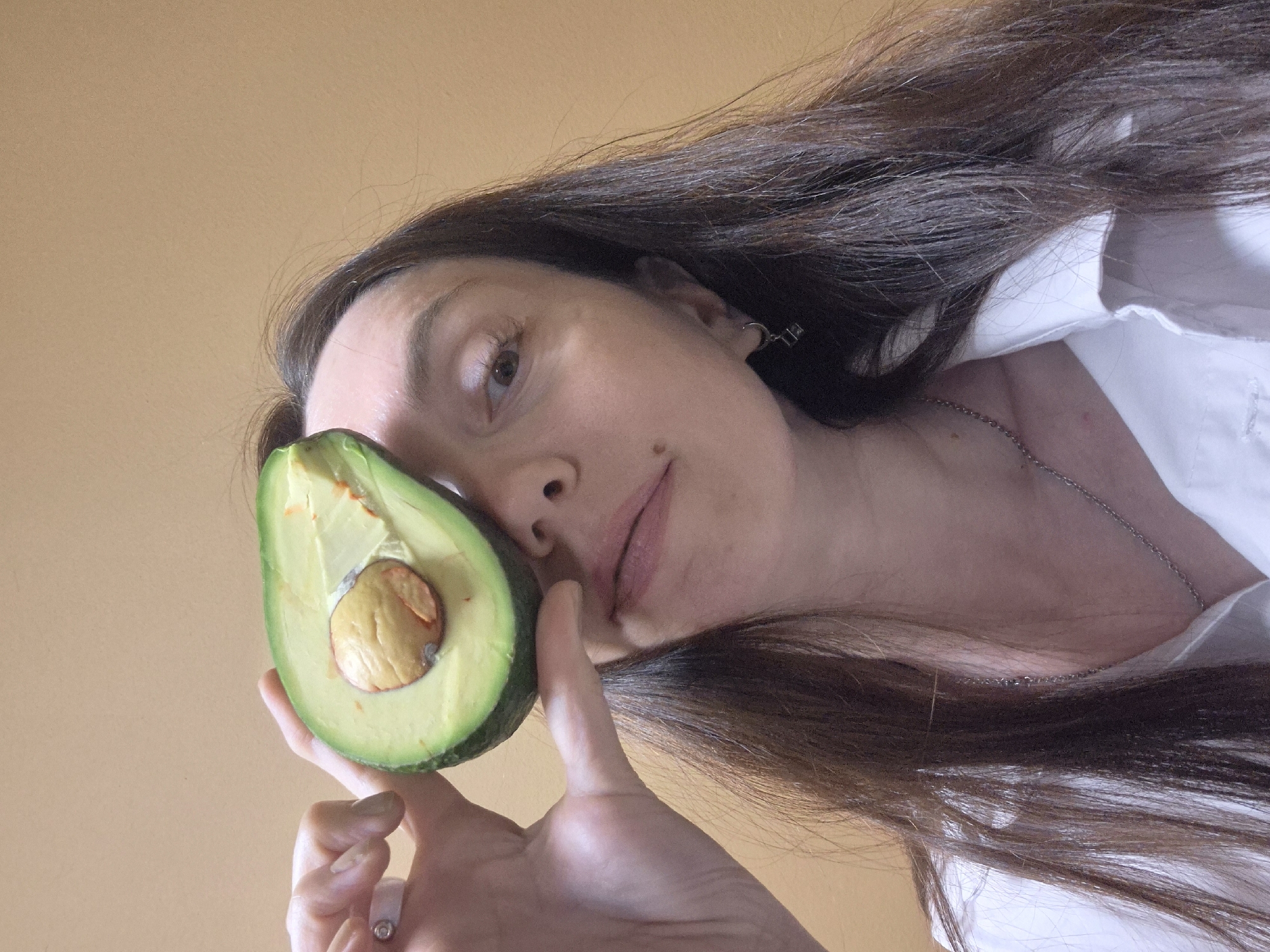 A woman with long, dark hair and fair skin is holding a halved avocado in front of her face. She is wearing a white collared shirt and a delicate silver necklace. Her left hand is visible, holding the avocado steady. She has a mole on her cheek. Her hair is loose and flowing around her face and shoulders. The background is a plain, light brown color. The photograph appears to be taken from a low angle, looking up at the woman.