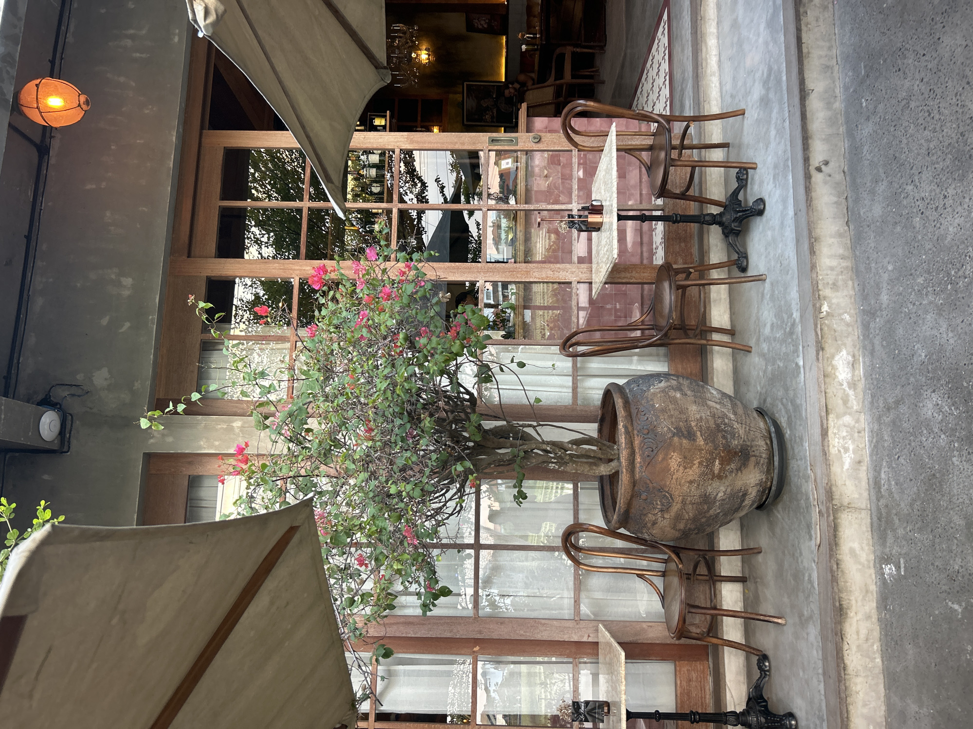 This image depicts the outdoor seating area of a restaurant in North Kuta, Indonesia. The scene features two tables with chairs, a large potted plant with pink flowers, and an umbrella providing shade. The restaurant's facade is visible, characterized by large wooden-framed windows and doors, with sheer curtains inside. On the right side of the image, a glimpse of the restaurant's interior reveals shelves stocked with bottles, a chandelier, and framed artwork. A patterned rug is visible on the floor inside. The overall impression is of a charming and well-maintained outdoor dining space.