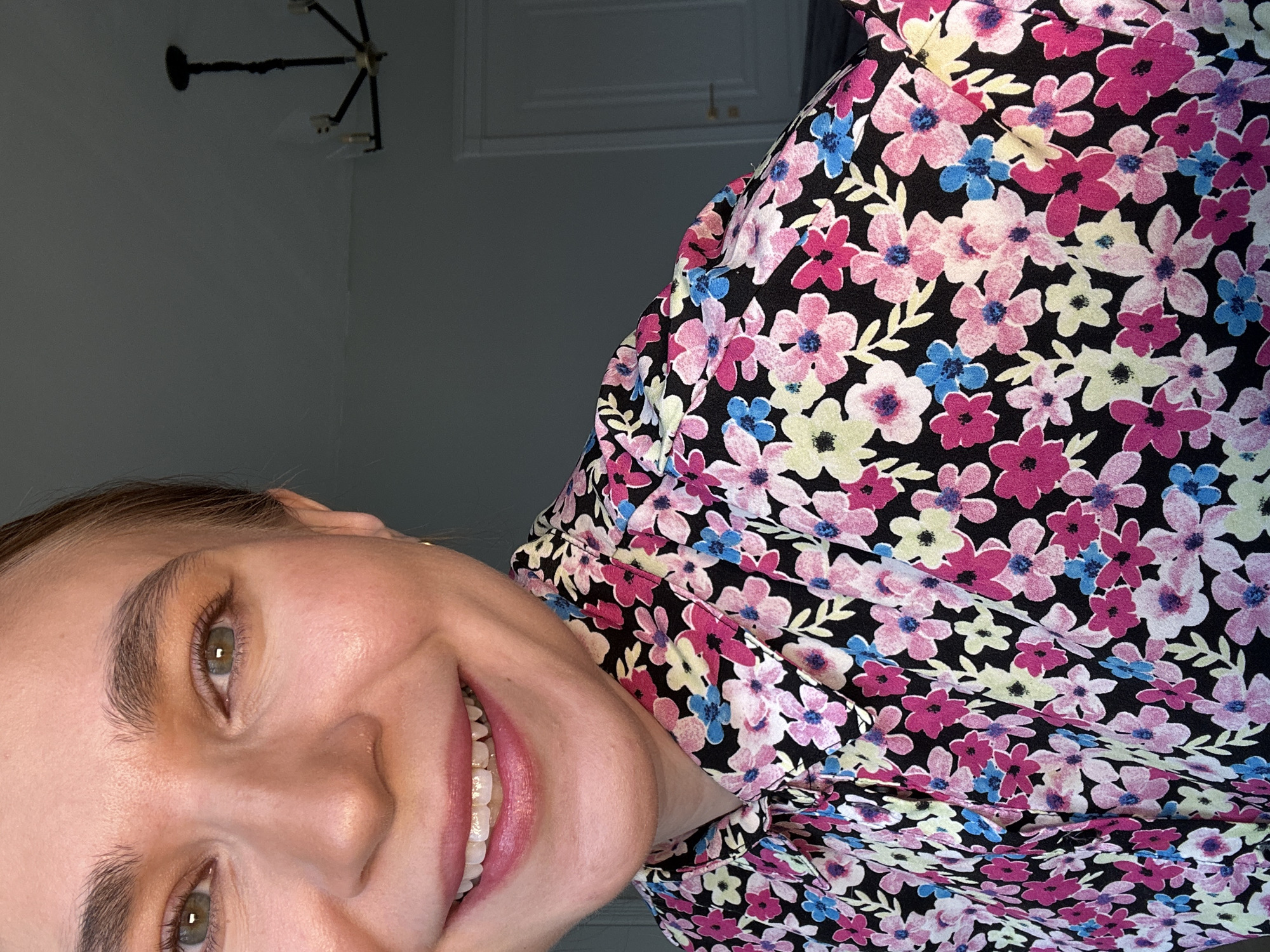 Here's a description of the image:
A close-up, tilted portrait of a woman with fair skin, light brown eyebrows, and light green eyes. She is smiling, showing her teeth. She is wearing a floral print dress with a black background and pink, blue, and yellow flowers. The background is a light gray wall with a white door and a black and gold light fixture. The location in the city of Kharnavy, Russia is not discernible from this image.