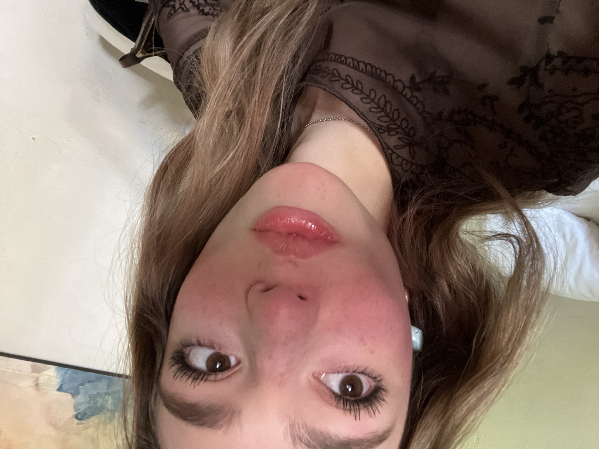 A close-up, upside-down selfie shows a young woman with fair skin, light brown hair, and brown eyes. She is wearing a brown lace top with a delicate floral pattern. Her lips are glossy, and she has a light dusting of freckles across her nose and cheeks. Her eyebrows are well-defined. A thin silver necklace is visible at her neck. In the background, a painting with blue and cream hues hangs on a wall. Part of a white pillow is also visible in the background. The location in the city of Puebla, Mexico, is not discernible from this image.
