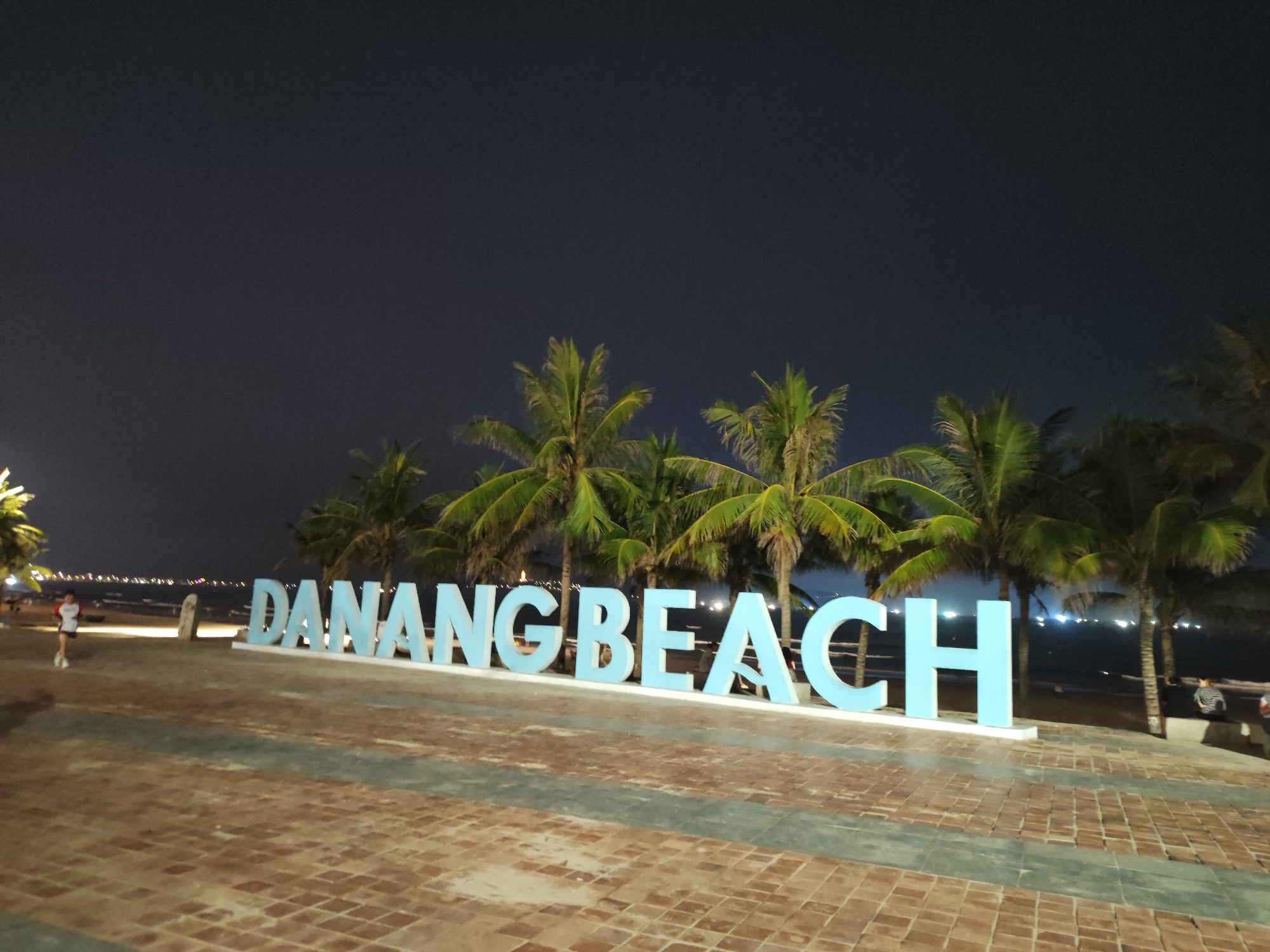 The image shows a nighttime scene at Đà Nẵng Beach in Vietnam. Large, light blue letters spelling "DANANG BEACH" are prominently displayed in the foreground. Behind the letters, there are several palm trees. The background includes the beach and the sea, with city lights visible in the distance. A person is walking on the left side of the image and people are sitting near the edge of the beach. The ground in the foreground is paved with bricks. The sky is dark, indicating it is night.