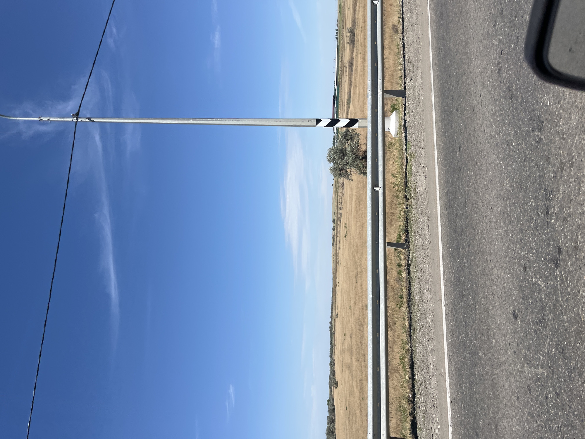 The image shows a roadside view in Bakhtemir, Russia, on a sunny day. A bright blue sky with wispy clouds stretches across the top of the frame, intersected by a power line. A tall, silver lamppost with black and white reflective bands stands prominently on the right side of the road. A silver guardrail runs along the side of the asphalt road, separating it from a dry, grassy field. In the field, a lone, bushy tree with light-colored foliage is visible, along with some distant structures and trees. The road surface in the foreground is asphalt, with a white line marking the edge of the lane. A portion of a car's side mirror is visible in the bottom right corner, suggesting the image was taken from inside a moving vehicle. The overall scene depicts a rural highway landscape.