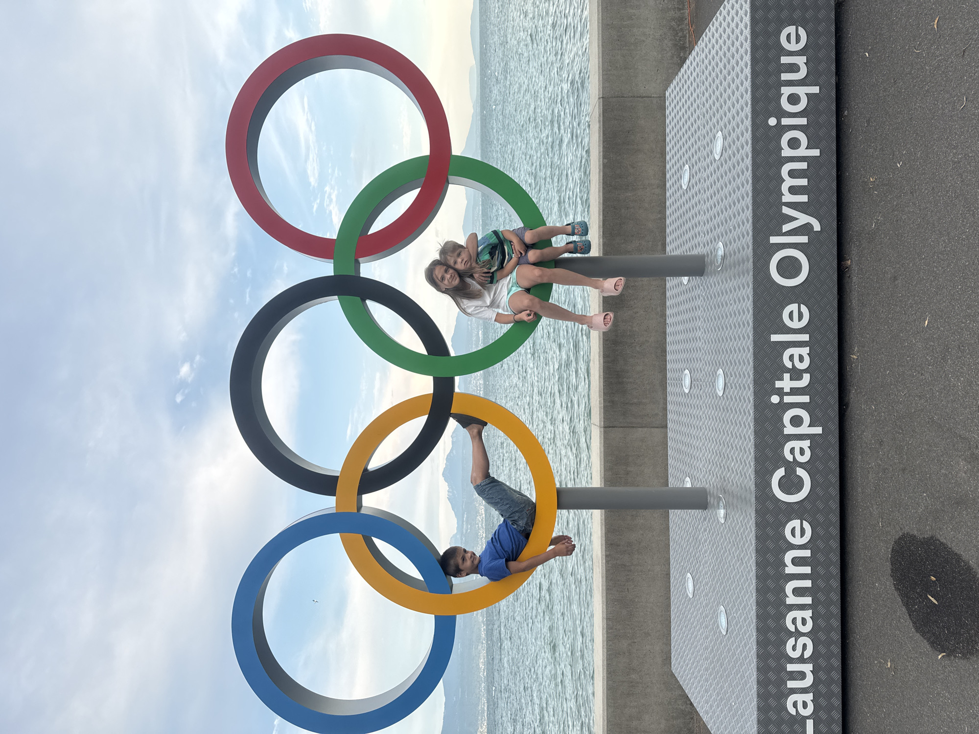 Two children and a boy are posing on a large Olympic rings sculpture located in Lausanne, Switzerland. The sculpture is situated along a waterfront promenade with a body of water in the background. The boy is sitting inside the yellow ring, with his legs extended. A girl and a younger boy are sitting together on the green ring, with the girl's arm around the younger boy. The Olympic rings are made of colorful metal and are supported by gray poles. The base of the sculpture has the words "Lausanne Capitale Olympique" written on it. The sky is cloudy and the water is calm.