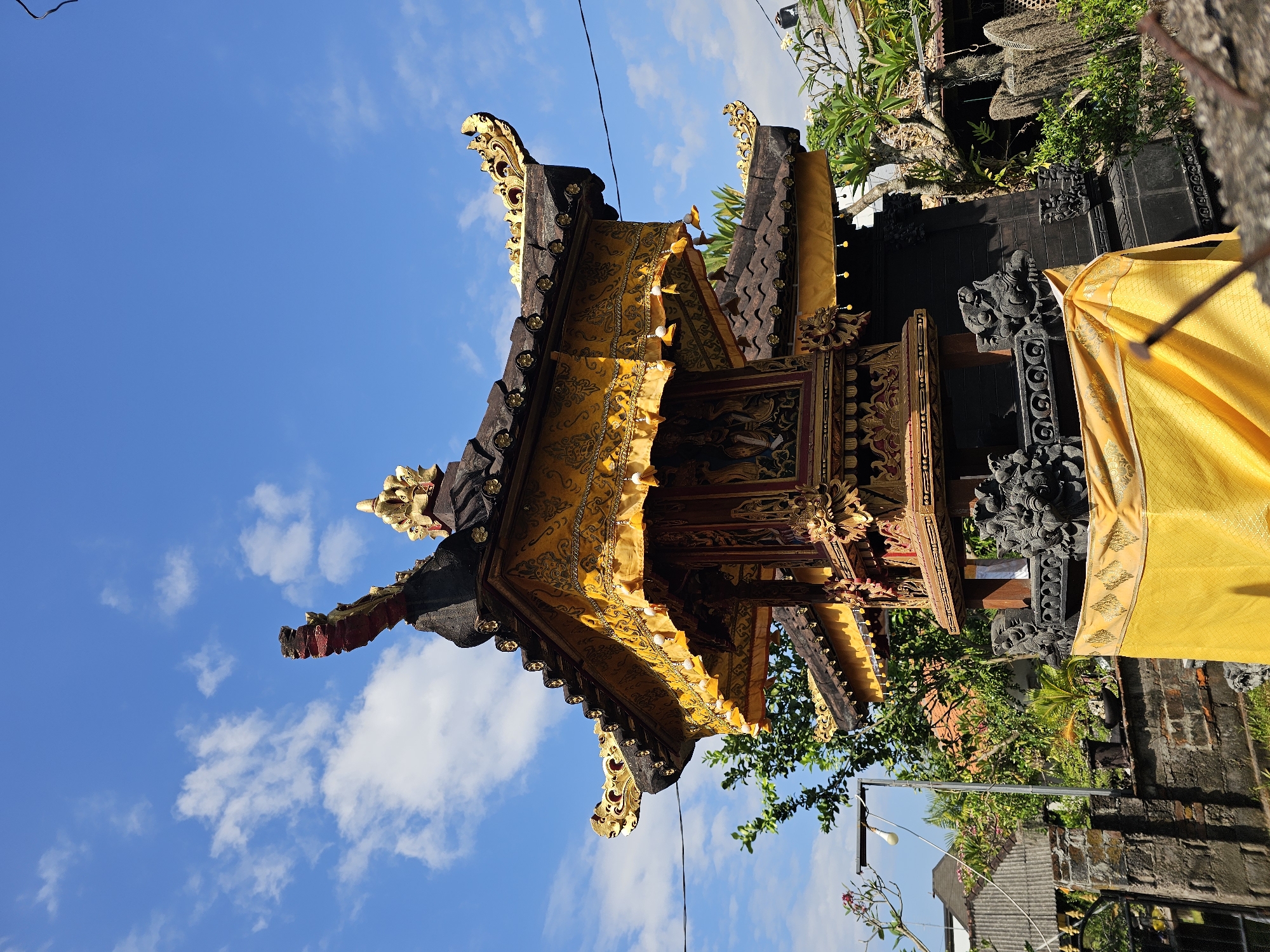 A Balinese temple is captured from a low angle against a bright blue sky with scattered white clouds. The temple structure features multiple tiered roofs with dark tiles and ornate golden finials. A yellow cloth with a decorative pattern and small white tassels drapes from the lower roof. The intricate details of the temple's woodwork, including carved figures and gold accents, are visible. In the foreground, a rough-textured wall is present, with a hint of green foliage and a light pole with a bulb visible to the left. The background reveals more greenery, rooftops, and a stone wall. The scene appears to be in a residential or temple complex, likely in Denpasar, Indonesia, given the architectural style and cultural context. There are no people or animals present in the image.