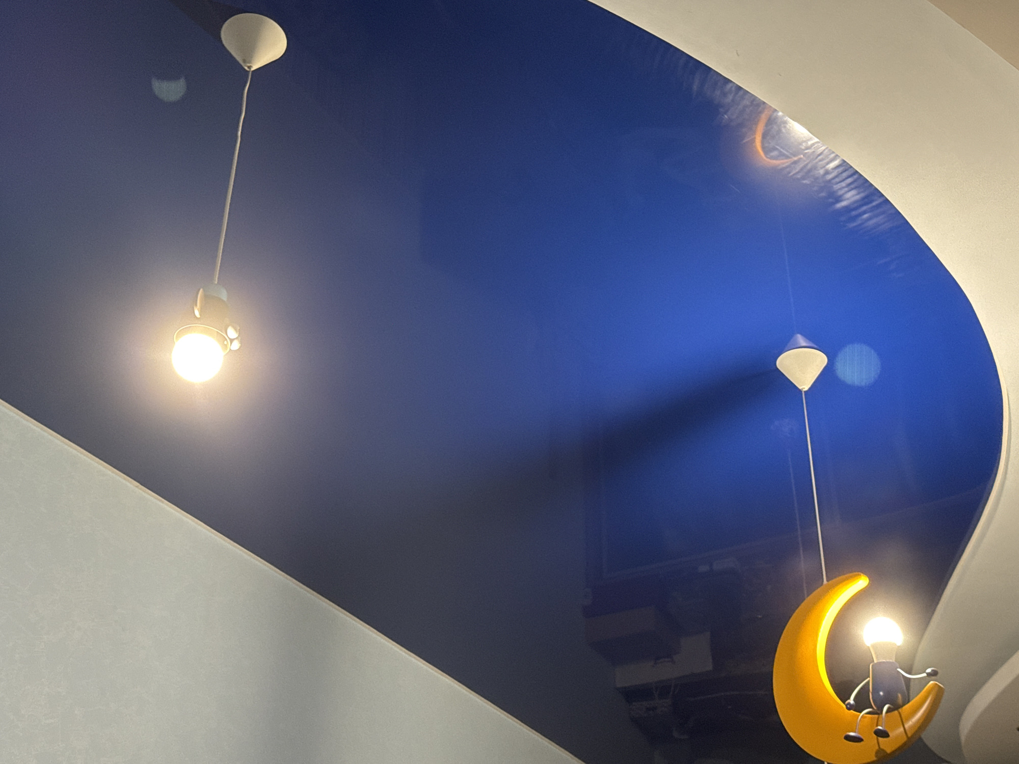 The image depicts a dimly lit indoor scene with a high, dark blue, reflective ceiling that appears to be a stretch ceiling or a lacquered surface. Two pendant lights are visible. The first light, on the left, is a simple bulb hanging from a white cord and a white cone-shaped fixture. It emits a warm, bright light. Further to the right and lower in the frame, a second, more elaborate light fixture is hanging. This fixture is shaped like a crescent moon, rendered in a bright yellow, and has a cartoonish figure sitting on it. This figure appears to be a small, dark blue body with white arms and legs, and a light bulb for a head. The light bulb head is illuminated, casting a soft glow. The ceiling reflects the lights and hints of objects on the floor or other surfaces above. The overall ambiance suggests a decorative lighting installation, possibly in a restaurant, bar, or themed room, given the whimsical moon light. The walls are a muted, light gray, angled, and partially visible. The scene is captured from a low angle, looking upwards. There are no people visible in the image. The location is unknown.
