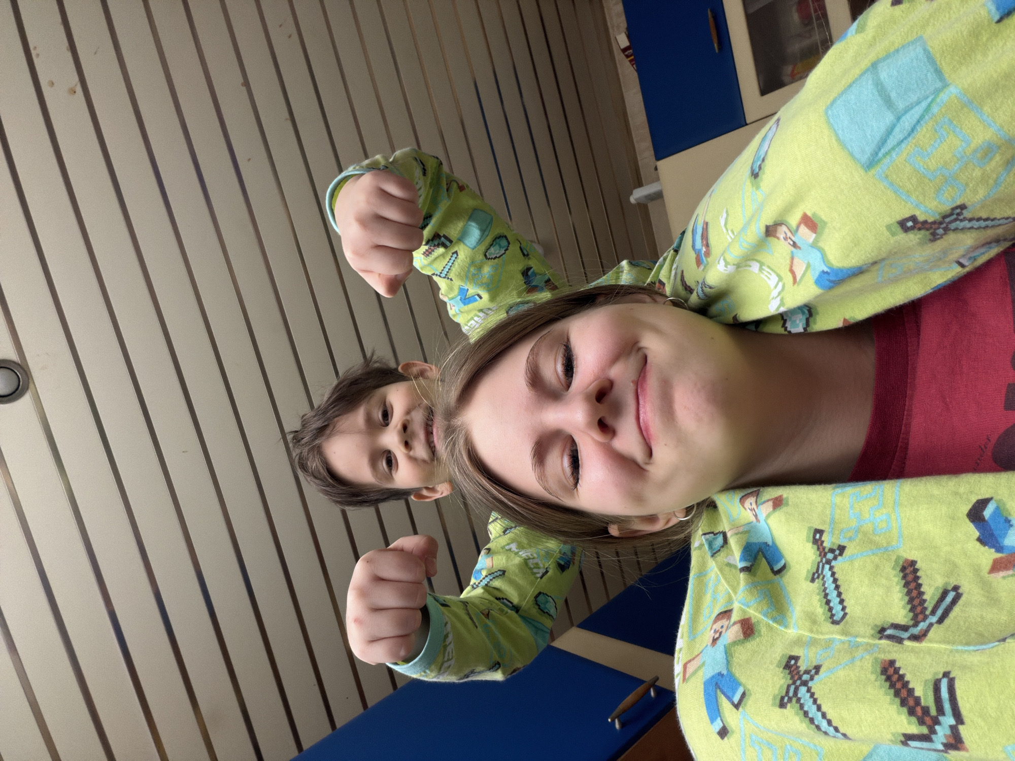Two people are in the image: a child and a young adult. The child is behind the young adult, appearing to hold their fists up. Both are wearing green pajamas with a Minecraft pattern. The young adult is smiling and looking into the camera. A portion of a blue surface is in the lower center. The background features a wall with vertical stripes. The environment appears to be indoors. The location, Perm, Russia is not recognizable based on the image content.