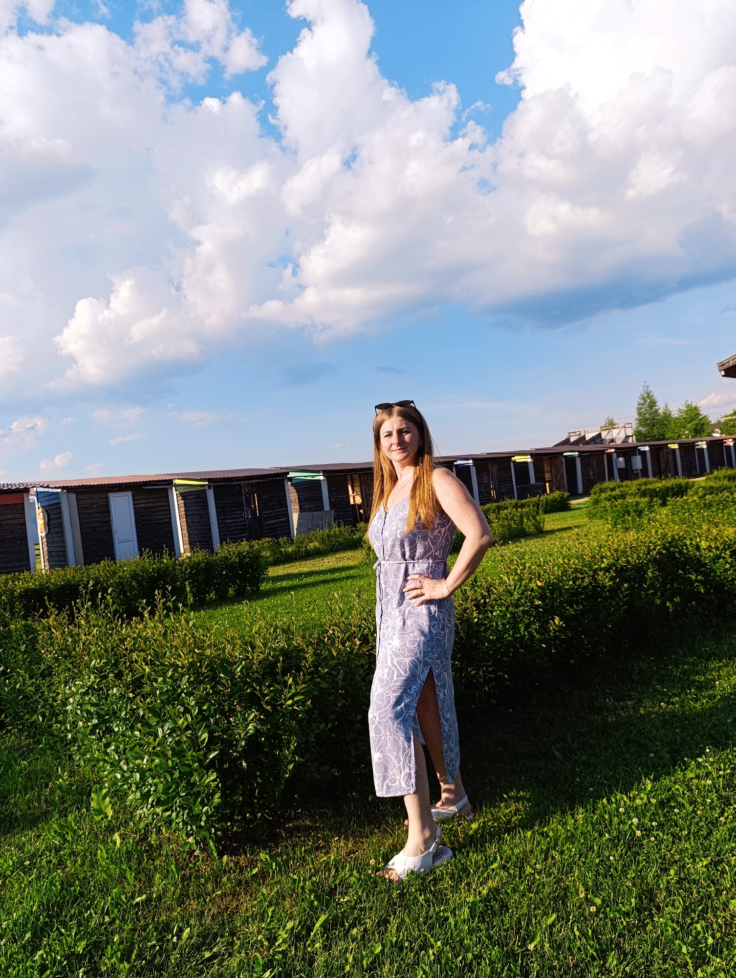 A full-body shot captures a woman standing on a grassy area with lush green bushes in the foreground and a row of small, wooden structures in the background. The sky above is a vibrant blue, dotted with fluffy white clouds. The woman, with long, light brown hair, is wearing a light blue dress with a white floral pattern and a thigh-high slit. She has on sunglasses pushed up on her head, a ring on her left hand, and is posing with her left hand on her hip. Her white sandals are visible on her feet. The structures in the background appear to be beach or changing huts, each with a dark wooden facade and a colorful trim along the roofline, suggesting a seaside or recreational location, possibly in Ivanovskoe, Russia. The lighting indicates it is daytime, likely late afternoon, with the sun casting shadows on the grass.