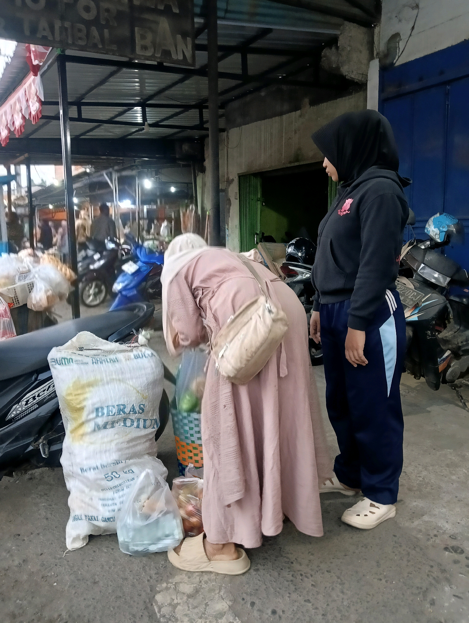 In Purwareja, Indonesia, two women are standing near a motorcycle. The woman on the right wears a black hijab and a black hoodie with a pink graphic, paired with dark blue track pants and beige slip-on shoes. She is facing left, looking down. The woman on the left is wearing a pale pink hijab and a pale pink long dress, also looking down. She has a beige crossbody bag slung over her right shoulder. In front of them, to the left, is a large white sack of rice labeled "BERAS MEDIUM" and "50 kg". To the right of the rice sack are two plastic bags, one containing what appears to be water and another containing produce like onions and tomatoes in a green and orange woven basket. A motorcycle is parked behind the rice sack, and another motorcycle is visible in the background. The scene appears to be an outdoor market or roadside stall, with a metal awning overhead and a blue door visible in the background. The ground is concrete.