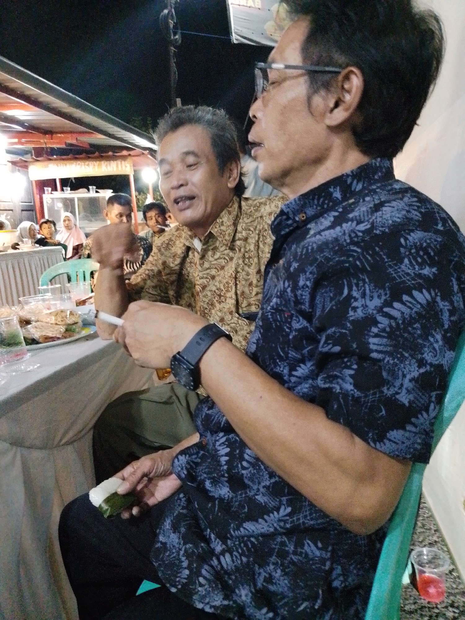A medium shot captures an outdoor gathering at night, likely in Trangkil, Indonesia, as suggested by the backdrop of a food stall with Indonesian text. Two men are the primary subjects. The man on the left, facing the camera in profile, is wearing a brown patterned shirt and appears to be speaking or laughing. In the foreground, a man in a dark blue patterned shirt faces right, holding a cigarette in his left hand and a wrapped food item in his right hand, which he is bringing towards his mouth. His left wrist is adorned with a black watch. In the background, other individuals, including women and children, are visible, some seated at tables. The environment suggests a casual dining or social event, with food items and drinks on the tables. The lighting indicates it is nighttime.