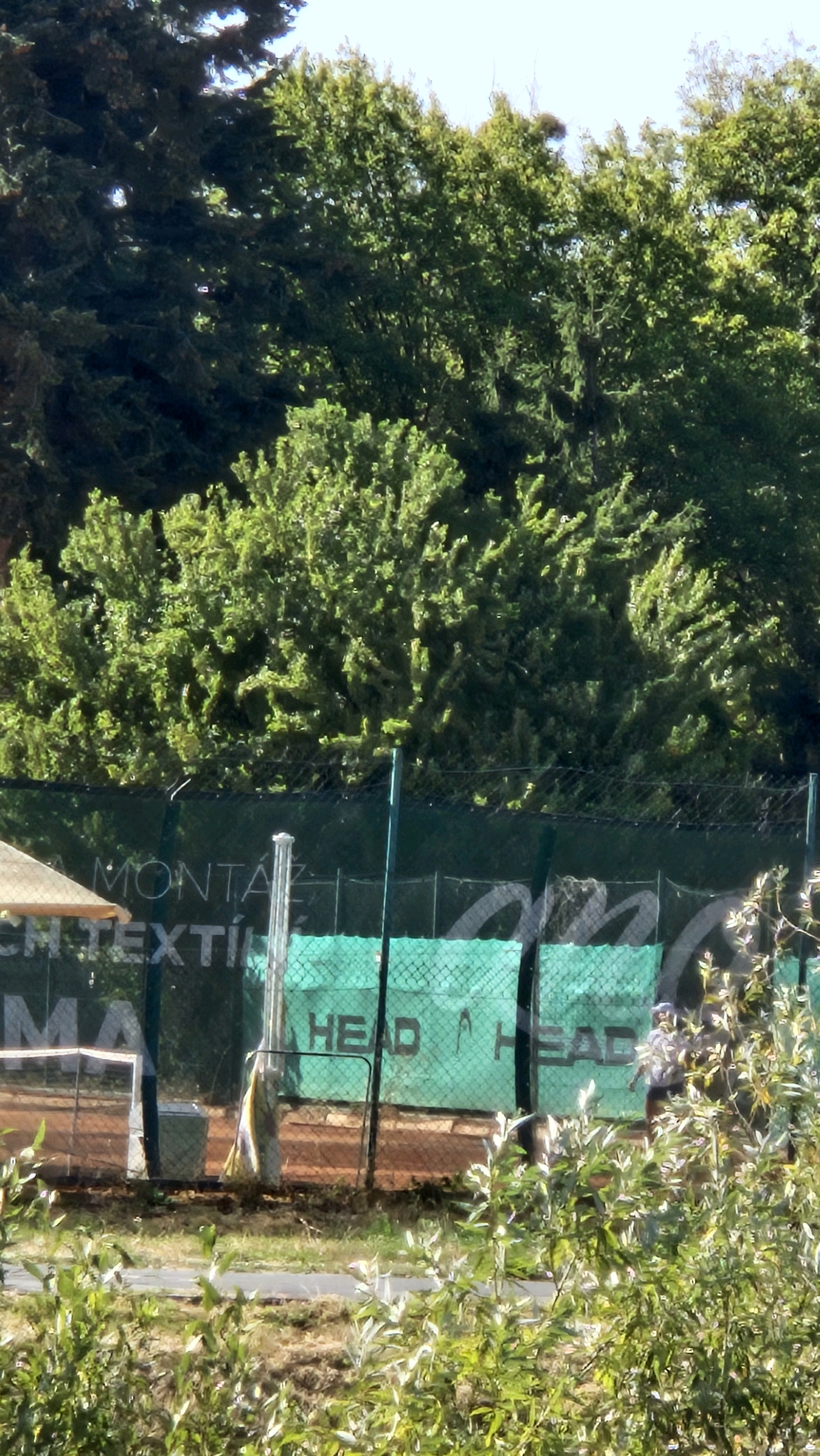 An outdoor tennis court in Prešov, Slovakia, is depicted, with a net partially visible in the foreground. The court is surrounded by a green fence with the "HEAD" logo printed on it. Trees and foliage frame the scene, with a clear sky overhead. A person can be seen in the background, appearing to be on the court. The ground of the court is brown, suggesting it is a clay court. A tan canopy is visible on the left side of the image, possibly providing shade.