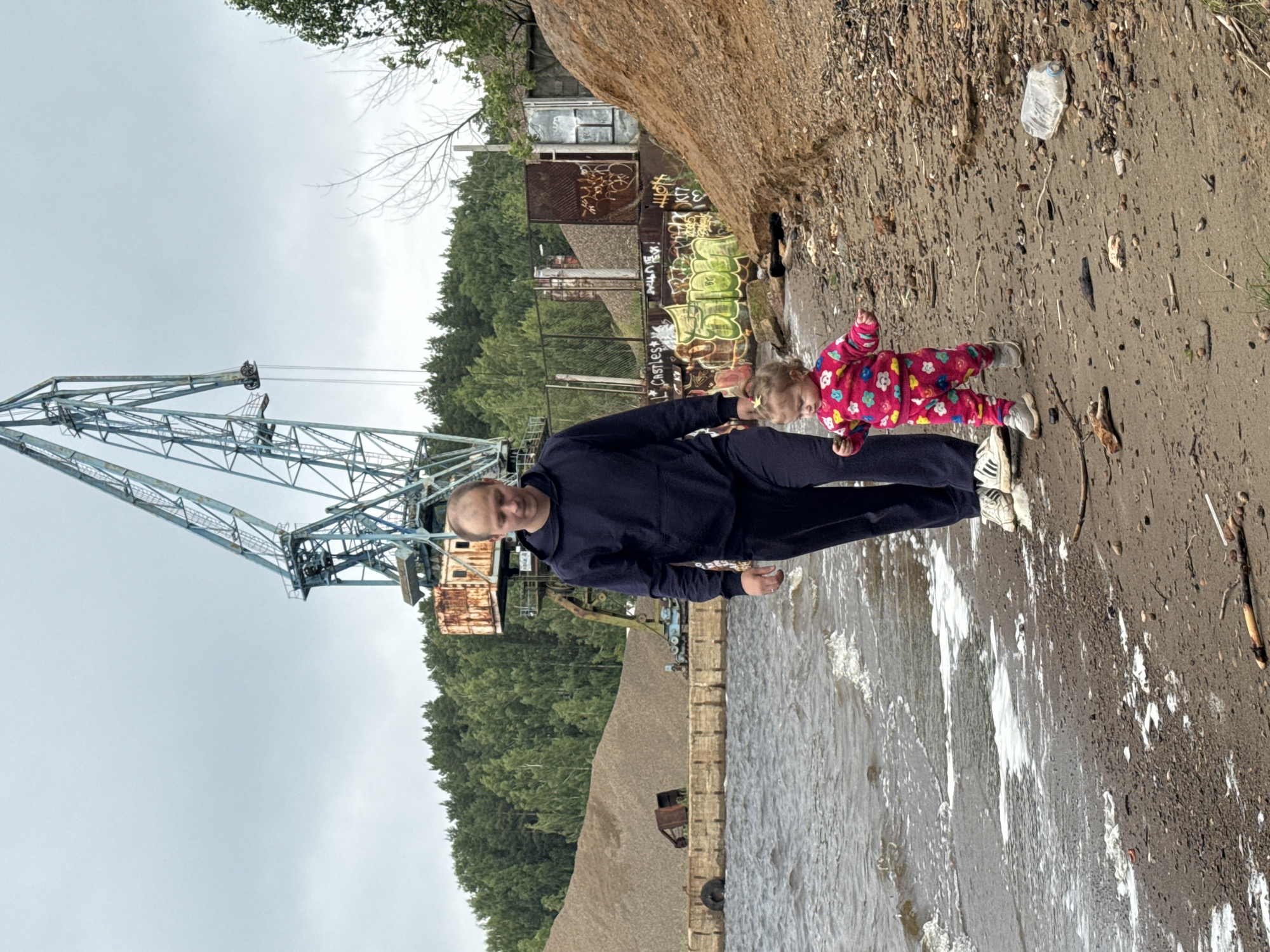 In this image, a man and a young child are standing near the water's edge. The man is wearing a dark blue hooded sweatshirt and dark pants, with white sneakers. The child is wearing a pink outfit with colorful floral patterns and white sneakers. They are positioned on a gritty, sandy shore with some debris, including a plastic bottle. The water appears to be choppy with some white foam.
In the background, a large industrial crane dominates the left side of the frame. It is a skeletal metal structure with visible cables and machinery. Behind the crane and to the right, there are trees and some dilapidated industrial buildings covered in graffiti. The graffiti includes visible text such as "3M YOT," "Castles," and other illegible tags. To the right of the people and the water, a large, textured boulder is prominent. The overall scene suggests an outdoor, possibly abandoned or industrial waterfront area. The sky is overcast, indicating a cloudy day. The time of day is not definitively ascertainable, but the lighting suggests it could be daytime. The atmosphere appears casual, with the man and child seemingly enjoying their time outdoors. The location is identified as Chaikovsky, Russia.