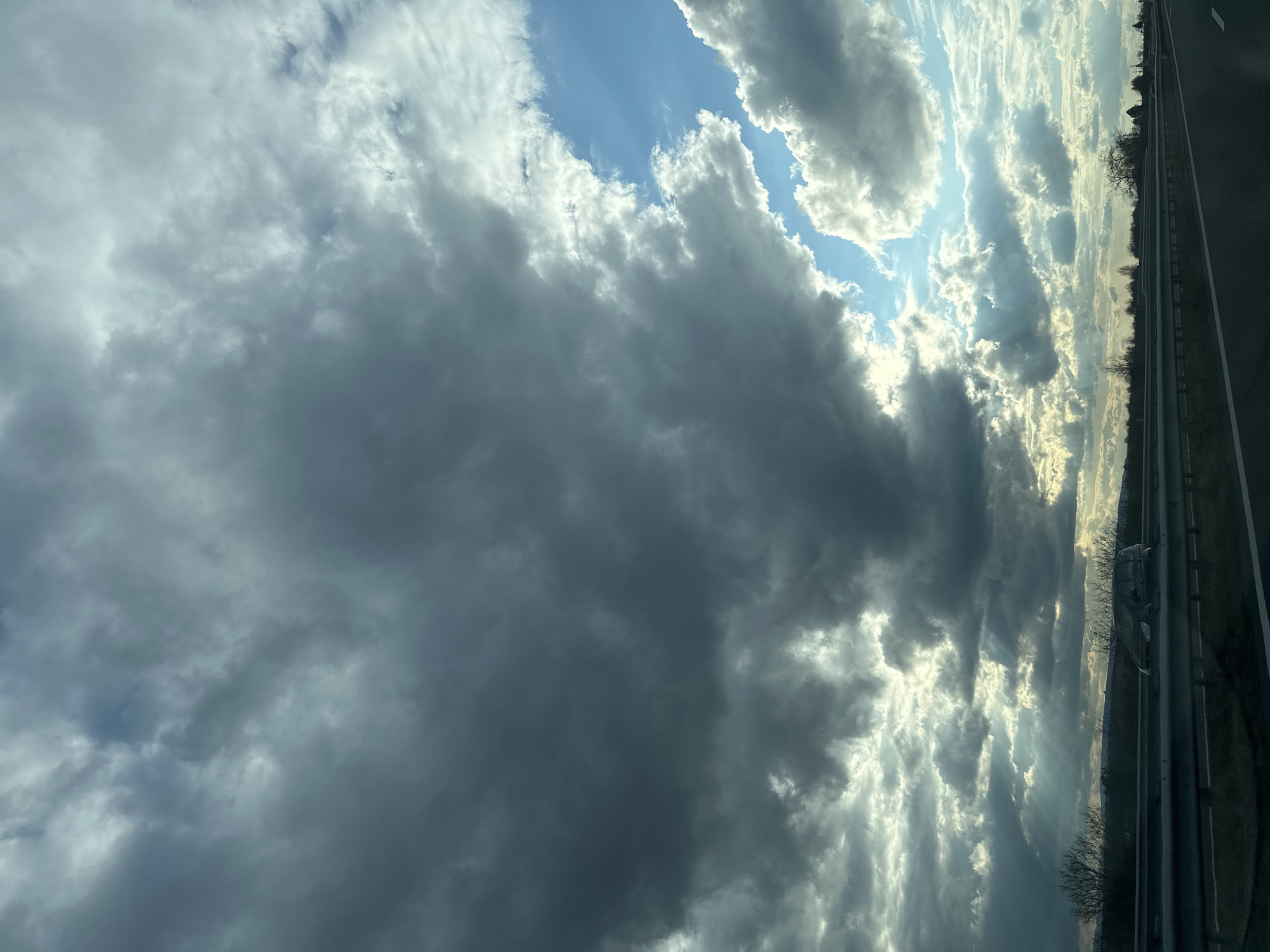 The image depicts a cloudy sky with patches of blue visible between the clouds. A road is visible on the right side of the frame, and there are trees and other vegetation along the road. The location is Kaspichan, Bulgaria.