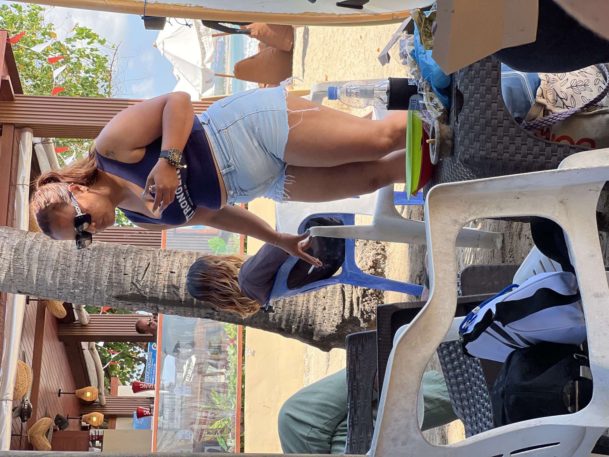 An outdoor scene unfolds in Kuta, Indonesia, likely at a beachside cafe. A woman, her dark hair pulled back in a ponytail and wearing sunglasses, stands and holds a cigarette and lighter. She is dressed in a dark blue tank top and light blue denim shorts. To her left, a person with shoulder-length, wavy hair sits in a blue plastic chair, facing away from the camera, and appears to be holding a cigarette. In the foreground, a white plastic chair is partially visible, with a black baseball cap and a white and blue bag resting on it. A woven plastic table in front of the chairs holds various items, including a green and red bowl, a small ashtray, a bottle of water in a koozie, and bags of snacks. Beyond the table, a palm tree trunk stands prominently, and a blurred background reveals beach umbrellas, beanbag chairs, and the ocean. The overall setting suggests a casual, relaxed atmosphere on a sunny day.