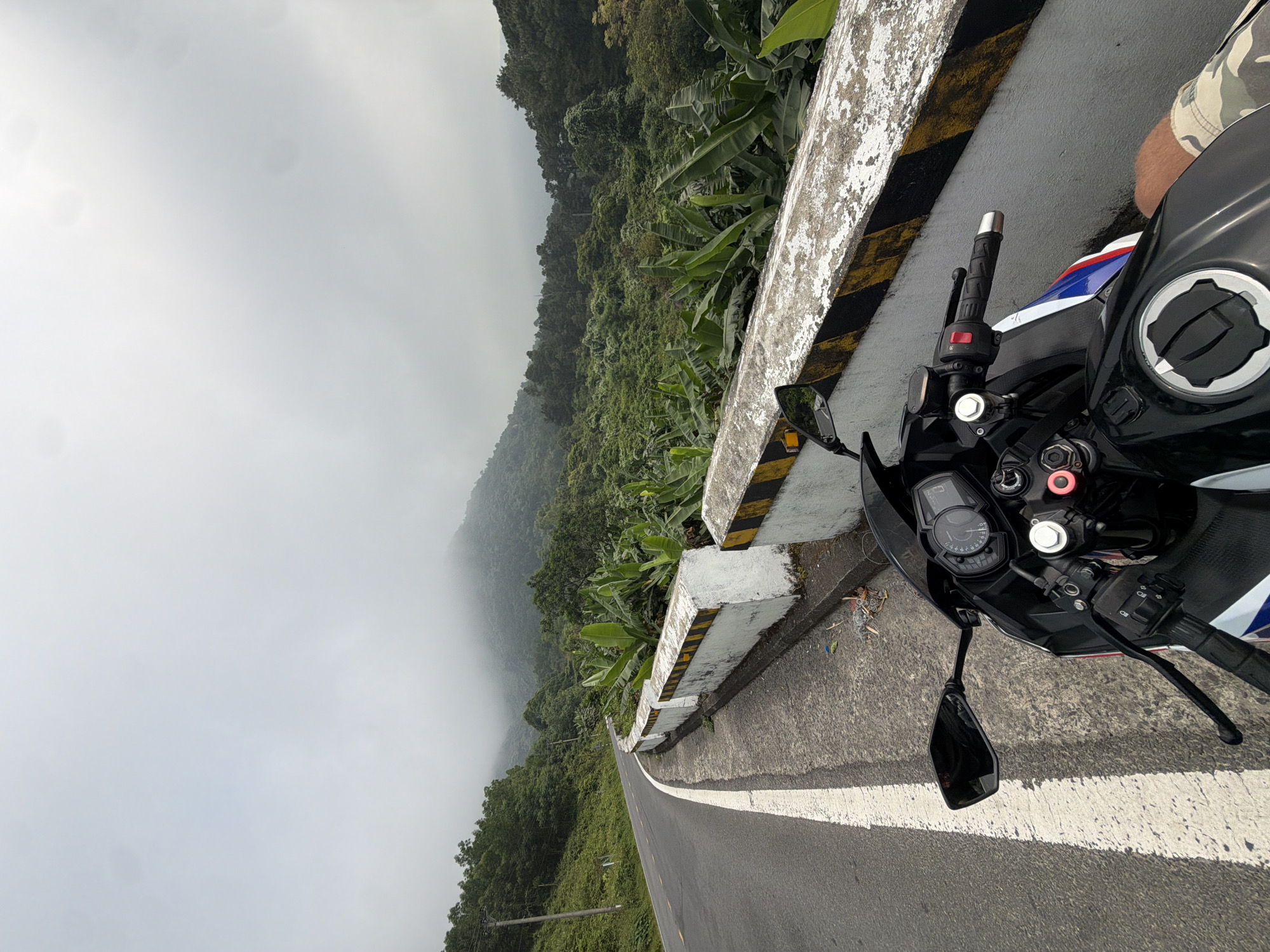 A motorcycle is parked on a winding road, possibly the Hai Van Pass, near Đà Nẵng, Vietnam. The weather is overcast, and fog obscures the view of the forested mountains. A concrete barrier, marked with alternating black and yellow stripes, runs along the edge of the road. The road itself is asphalt with a white dividing line. The motorcycle's handlebars, instrument panel, and a portion of the tank are visible. A person wearing camouflage clothing is partially visible on the right side of the frame.