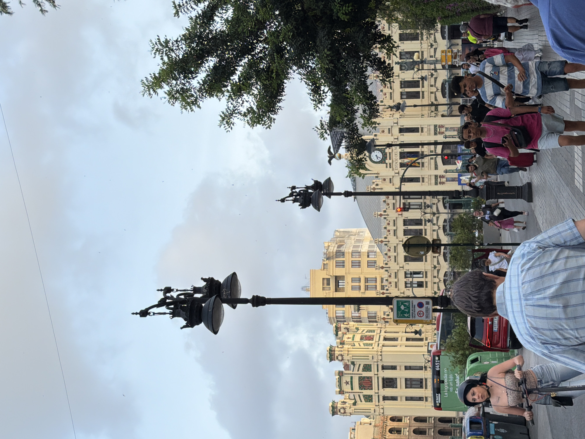 In a bustling city square, two ornate street lamps stand tall against a cloudy sky, their intricate designs a contrast to the modern green bus and red car parked nearby. A young woman with a helmet rides a scooter, her gaze fixed forward, while a man in a plaid shirt walks away from the camera. A clock tower, adorned with an eagle sculpture, overlooks the scene from a historic building. A crowd of people navigates the pedestrian walkway, some on foot, others with luggage, amidst a backdrop of architectural grandeur. The atmosphere is lively, with a sense of movement and urban activity.