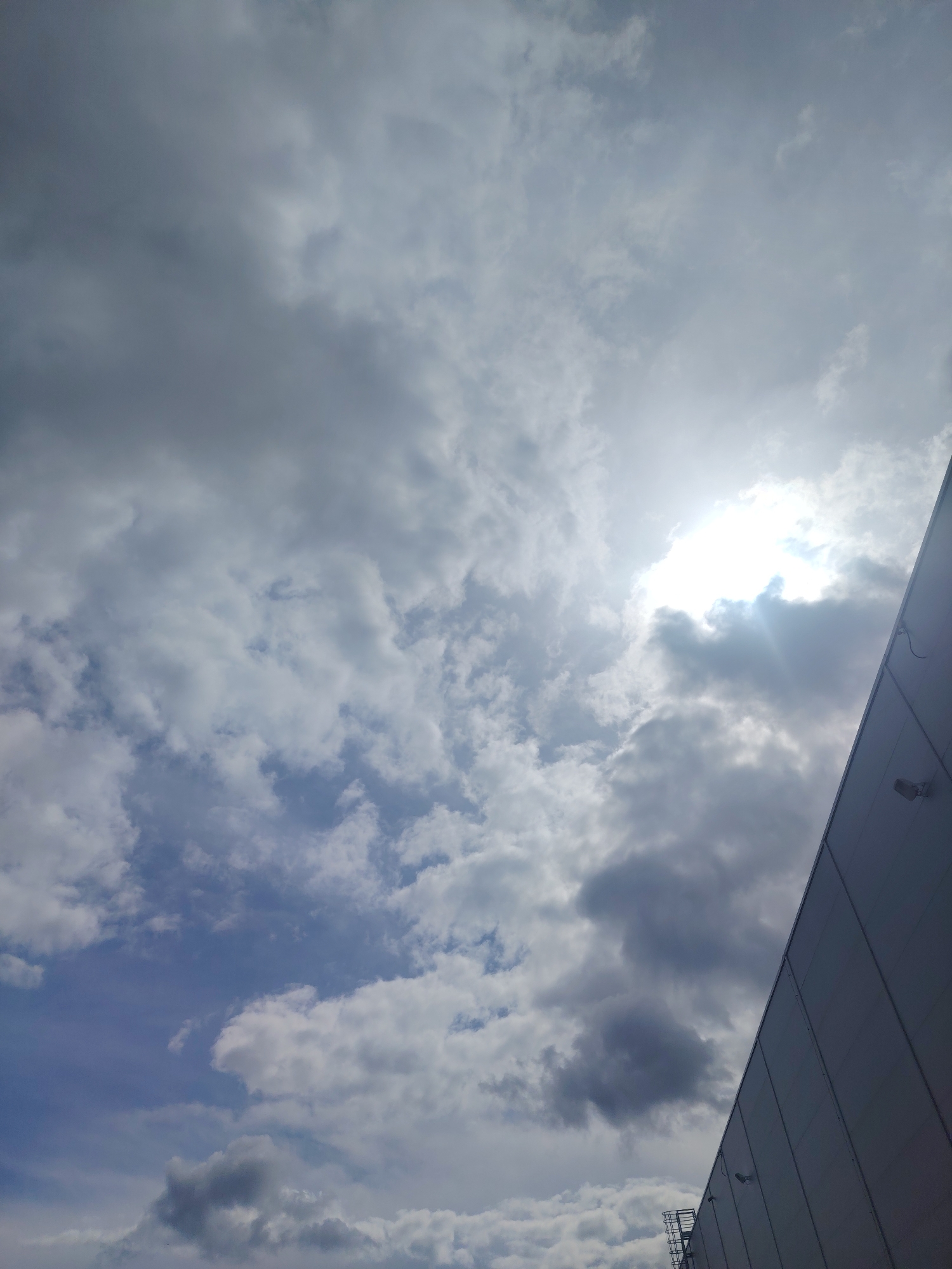 The image shows a sky filled with clouds. The sun is visible, peeking through some of the clouds, creating a bright glare. In the lower right portion of the image, a section of a building's exterior is visible, appearing to be a large, modern structure with a light-colored, possibly metallic facade. There are a few fixtures, resembling lights or cameras, mounted on the building. A metal ladder or stairway structure is also visible at the bottom edge of the building. The scene suggests an outdoor environment, possibly in a commercial or industrial area given the building's appearance. The weather appears overcast with patches of sunlight.