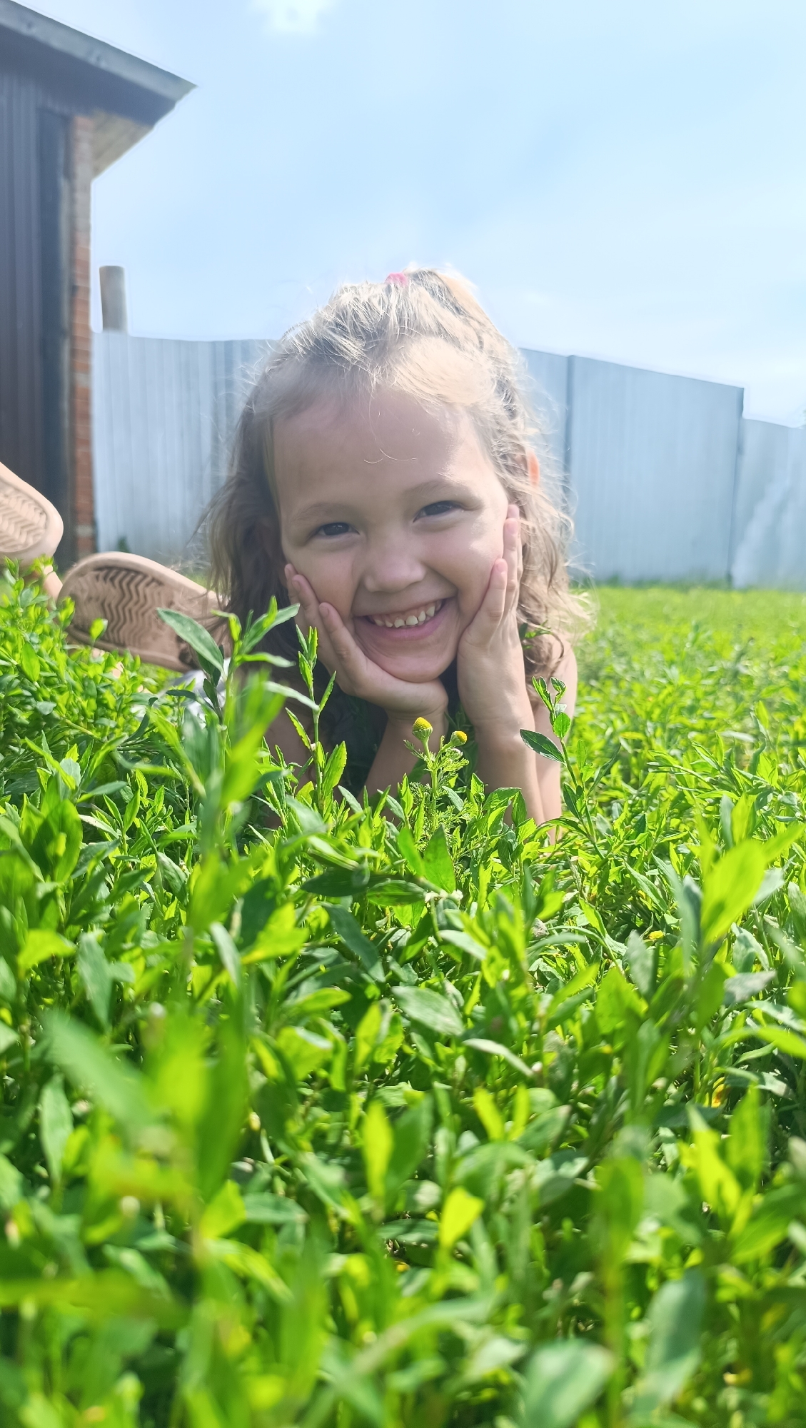 In Karsha, Russia, a young girl with blonde hair pulled into a ponytail, wearing a green top, lies on her stomach in lush green grass. Her face is turned towards the camera, and she is smiling broadly, with her hands supporting her chin. In the background, a corrugated metal fence and a brick building are visible under a bright sky with scattered clouds. The lower half of a sandal is also visible on the left side of the frame. The focus is on the girl and the vibrant green grass in the foreground.