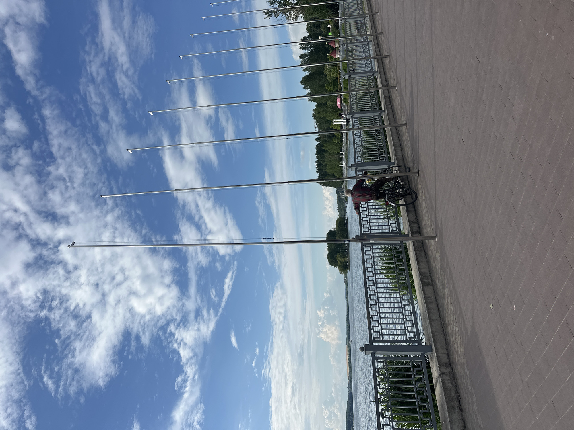 The image shows a man on a bicycle facing away from the camera, leaning on a railing. He is wearing a plaid shirt and dark pants. The bicycle is black. Behind him is a body of water, likely a lake or river, with a shoreline in the distance featuring trees and some buildings. A series of tall, thin metal poles or flagpoles line the promenade in front of the railing. The promenade itself is paved with brick-like stones. The sky is blue with scattered white clouds. The scene is set in Ternopil, Ukraine, likely on the promenade of Ternopil Lake, a popular recreational area.