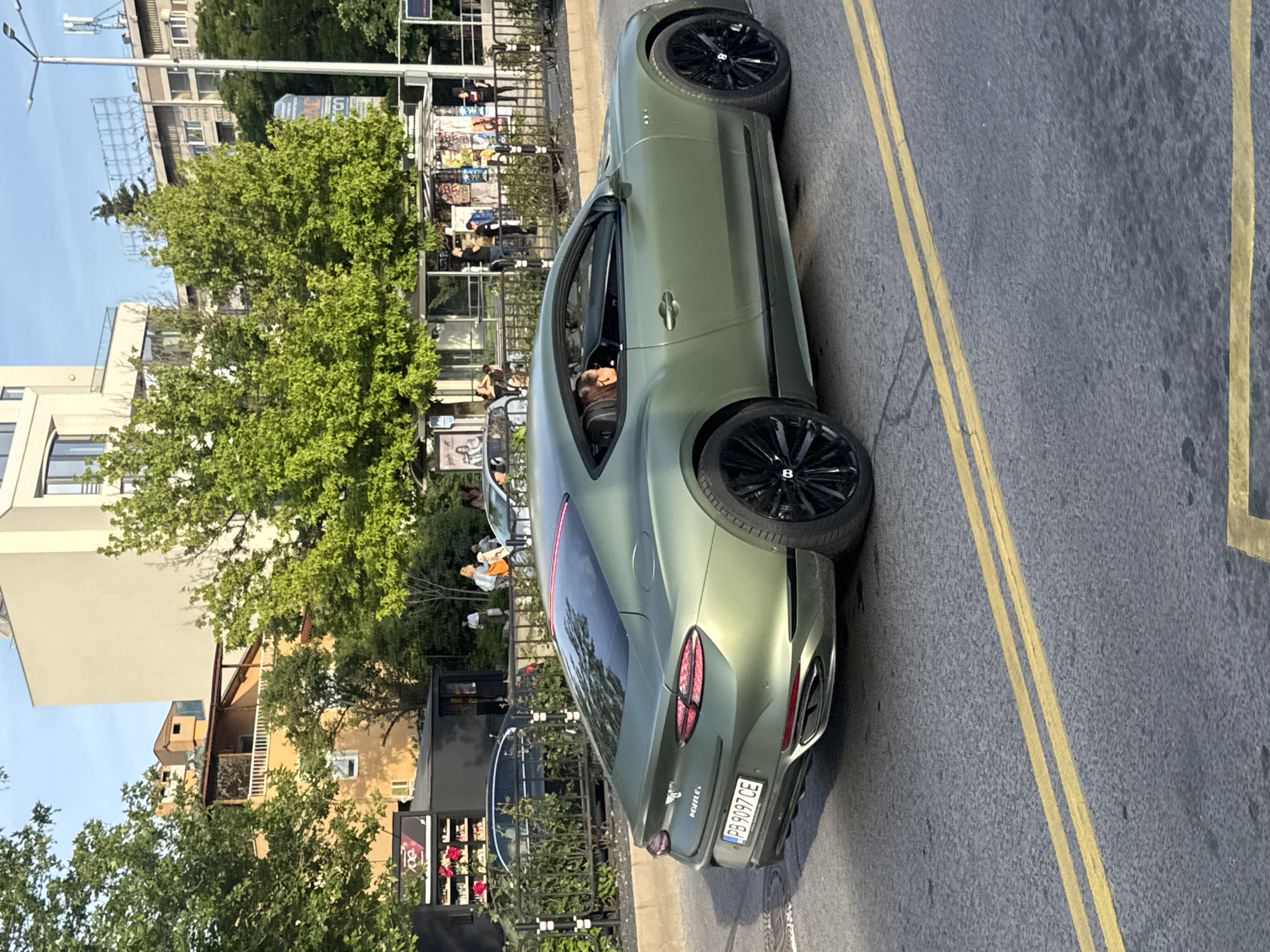 A green Bentley Continental is parked on the side of the road near a bus stop in Plovdiv, Bulgaria. The car is facing away from the camera. The license plate number is PB 9097 CE. The car is parked on a gray asphalt road with double yellow lines. A person can be seen in the driver's seat. A group of people are standing near the bus stop, which is near a black metal fence. Trees and buildings are visible in the background. The weather appears to be sunny.