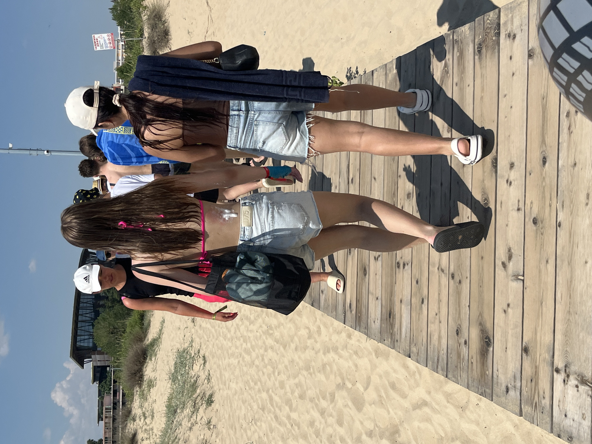 The media file captures a group of people walking along a wooden boardwalk on a sunny day at a beach. The setting appears to be a resort area, with sandy ground and sparse vegetation bordering the boardwalk. In the background, a modern building complex with glass windows and an elevated structure is visible. There are also some distant trees and what appear to be smaller structures or ruins further in the distance.
The people in the foreground are casually dressed for a warm day, with two young women prominently featured. One is wearing denim shorts and a pink bikini top, and the other is also in denim shorts. Another individual, wearing a white cap and a black t-shirt, is visible to the left, and appears to be looking towards the camera. The overall mood seems relaxed and summery. The sun is high in the sky, casting distinct shadows on the wooden planks, suggesting it is likely midday.
One of the individuals is carrying a black mesh bag. There is a visible sign in the background with text in what appears to be Bulgarian, including hazard symbols and possibly information regarding beach rules or local attractions. The scene conveys a typical beach day experience with people enjoying the outdoors.