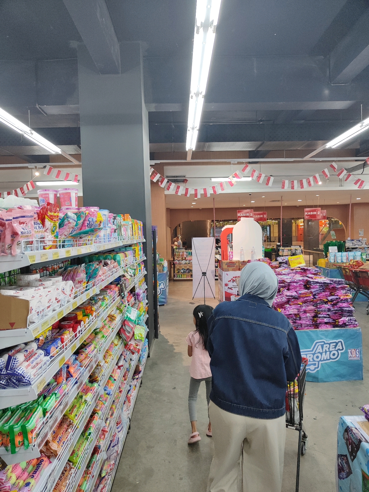 The image depicts an interior view of a brightly lit supermarket, possibly located in Sukowiryo, Indonesia, as suggested by the context. The scene is dominated by rows of well-stocked shelves filled with various consumer goods, predominantly packaged food items and household products. Red and white checkered flags are strung across the ceiling in a festive manner, indicating a possible national celebration or promotional event.
In the foreground, two individuals are visible. A woman wearing a grey hijab and a blue denim jacket is pushing a shopping cart, her back to the camera. Beside her and slightly ahead, a young girl in a pink shirt and grey pants is walking. Their presence suggests a shopping expedition.
Further into the store, several cashier stations are marked with signs "KASIR 1," "KASIR 3," and "KASIR 2/5." To the right of the woman, a large display of pink packaged items, possibly clothing or textiles, is arranged, along with a blue promotional banner that reads "AREA PROMO KDS." The overall environment suggests a busy retail space with merchandise neatly organized on shelves and promotional displays strategically placed. The floor is a light gray concrete, reflecting the overhead lighting. The ceiling is dark gray, with prominent exposed beams and fluorescent light fixtures illuminating the aisles.