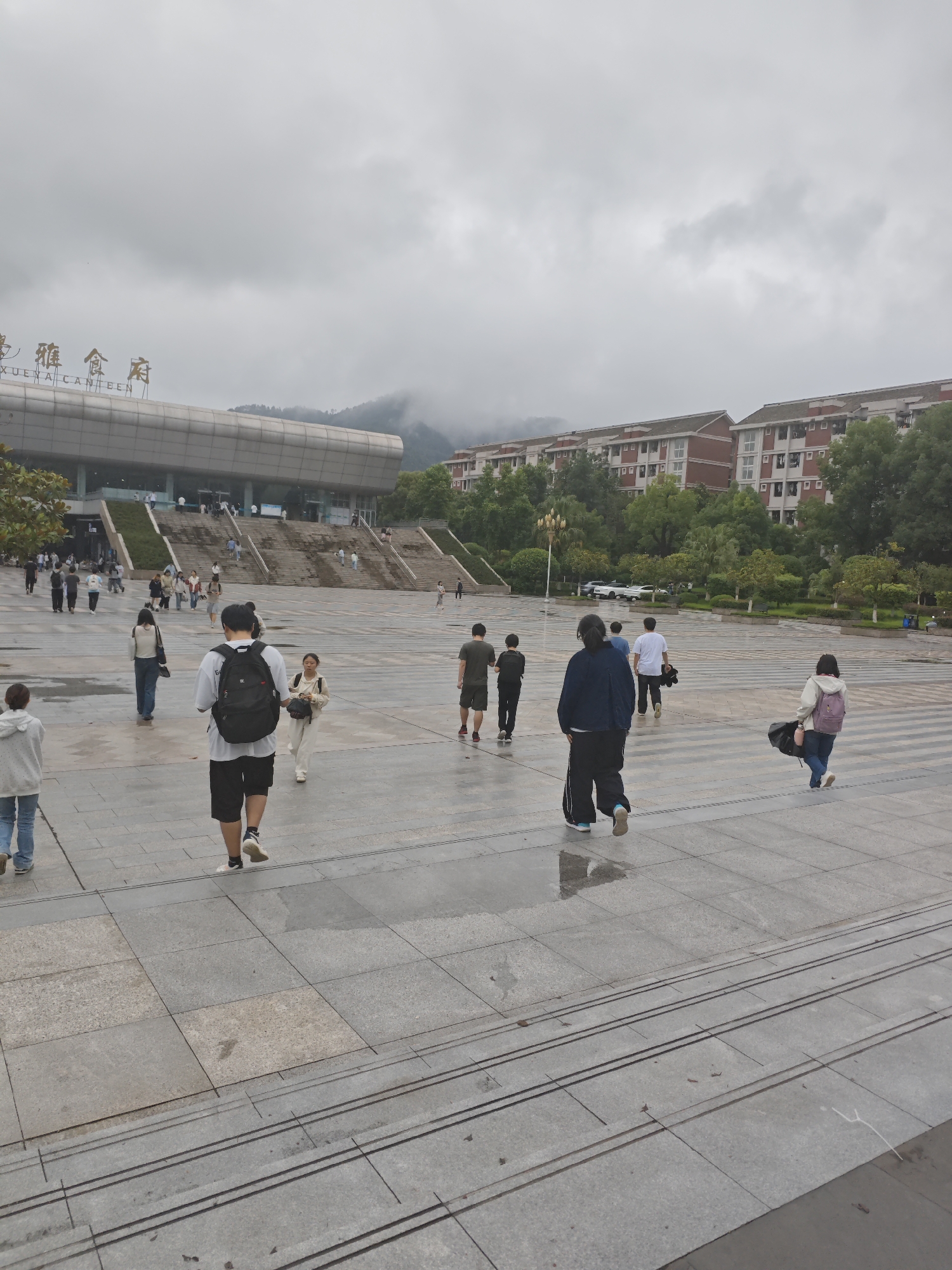 The image shows a wide, wet, outdoor plaza with a large, modern building in the background, identified by signage as "XUANA CANTEEN". The building has a metallic roof and is accessed by a wide set of stairs. To the right of the canteen, there are several multi-story residential buildings with red brick facades and white window frames, surrounded by lush green trees. The plaza itself is paved with large, square grey tiles, many of which are wet and reflective, indicating recent rain. There are numerous people walking across the plaza in various directions. Some are walking alone, while others appear to be in small groups. Many are carrying backpacks. The sky is overcast with heavy grey clouds, and the overall lighting suggests a gloomy, possibly humid, atmosphere typical of Chongqing. The ground reflects the sky and surrounding structures, further emphasizing the wetness of the plaza. The scene appears to be a public space on a university campus or a similar institutional setting, likely within the Chongqing High-tech Zone, China, given the architectural style and presence of a canteen and residential buildings.