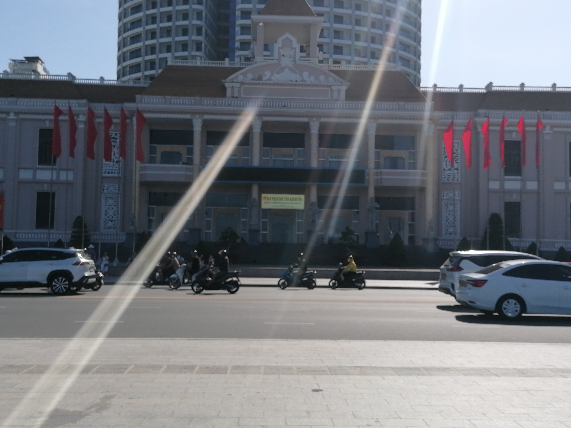 The image shows a street scene in Nha Trang Ward, Vietnam, with a large government building in the background. The building has a light grey facade, with a brown roof and red flags hanging from the front. A tall, modern building is visible behind the government building. In the foreground, there is a street with cars and motorbikes driving on it. The street is made of grey pavers. The sky is bright and clear. There are a few people visible on the sidewalks and on the motorbikes.