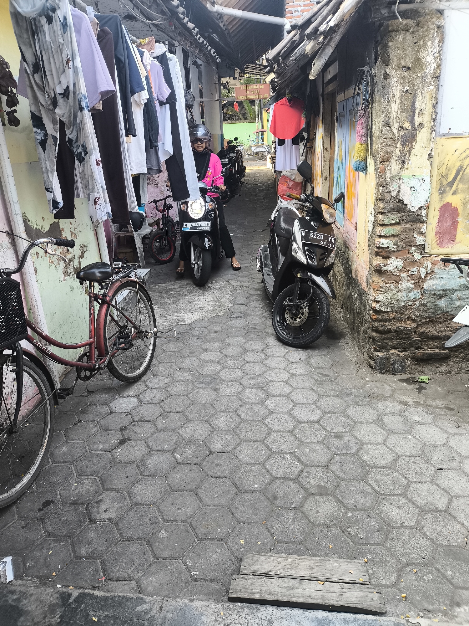 In a narrow alleyway in Citrodiwangsan, Indonesia, a woman wearing a pink jacket and a black helmet is seated on a black scooter. The scooter, with license plate N 6220 YR, is positioned in the center of the alley, facing forward. To the left of the scooter, a red bicycle with a basket is parked. The alleyway is paved with hexagonal grey stones. Clothes are hanging on a line overhead, obscuring the view of buildings. The walls of the buildings are painted in various colors, including yellow, pink, and blue, and show signs of age and weathering. A second, black scooter is visible further down the alley on the right. A white motorcycle is partially visible on the far right edge of the image.
