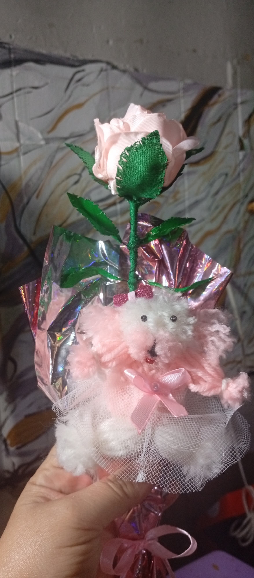 A hand holds a decorative bouquet with a pink rose and a pink and white stuffed animal. The bouquet is wrapped in shiny pink foil. The background appears to be a textured wall. There is no information available to confirm the location in the city of Juan Griego, Venezuela.