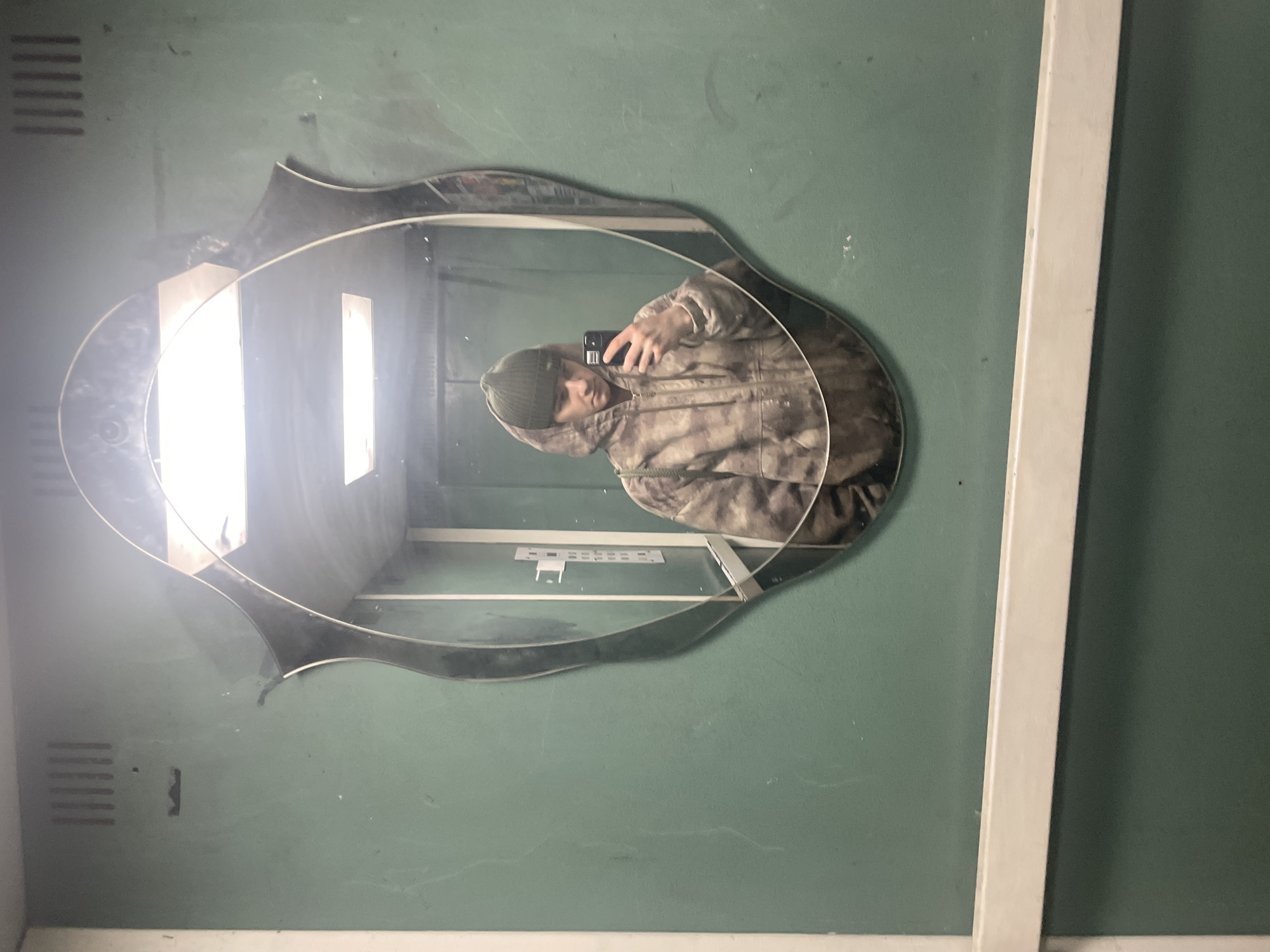 A person wearing a camouflage hoodie and a green beanie is taking a selfie in a decorative mirror. The mirror is hanging on a green wall in what appears to be a public restroom or changing room. There are horizontal vents visible on the wall. The mirror reflects the interior of the space, showing a white interior with vertical lighting. The location is presumed to be in Krasnoyarsk, Russia, based on user specification.