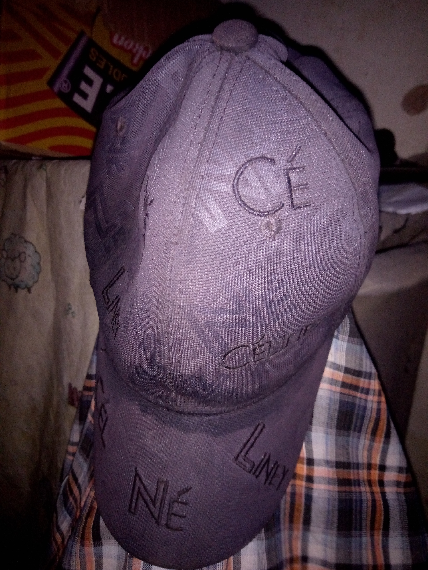 The image displays a dark purple baseball cap with repeated dark purple lettering. The letters "CE" are visible on the front of the cap, and "LINE" and "NE" are also present in a repeating pattern across the cap. The cap is resting on a checkered fabric, which appears to be a shirt or blanket, featuring a plaid pattern of blue, white, and orange. In the background, to the left, a yellow and orange striped item with black text that reads "E. LE NO" is partially visible. To the left of the cap, a light-colored fabric with a cartoon drawing of a sheep is also visible. The lighting in the image is dim, casting shadows and making some details difficult to discern. The overall scene appears to be indoors, possibly in a domestic setting. There are no individuals present in the frame. The exact location in Jalingo, Nigeria cannot be determined from the visual information provided.