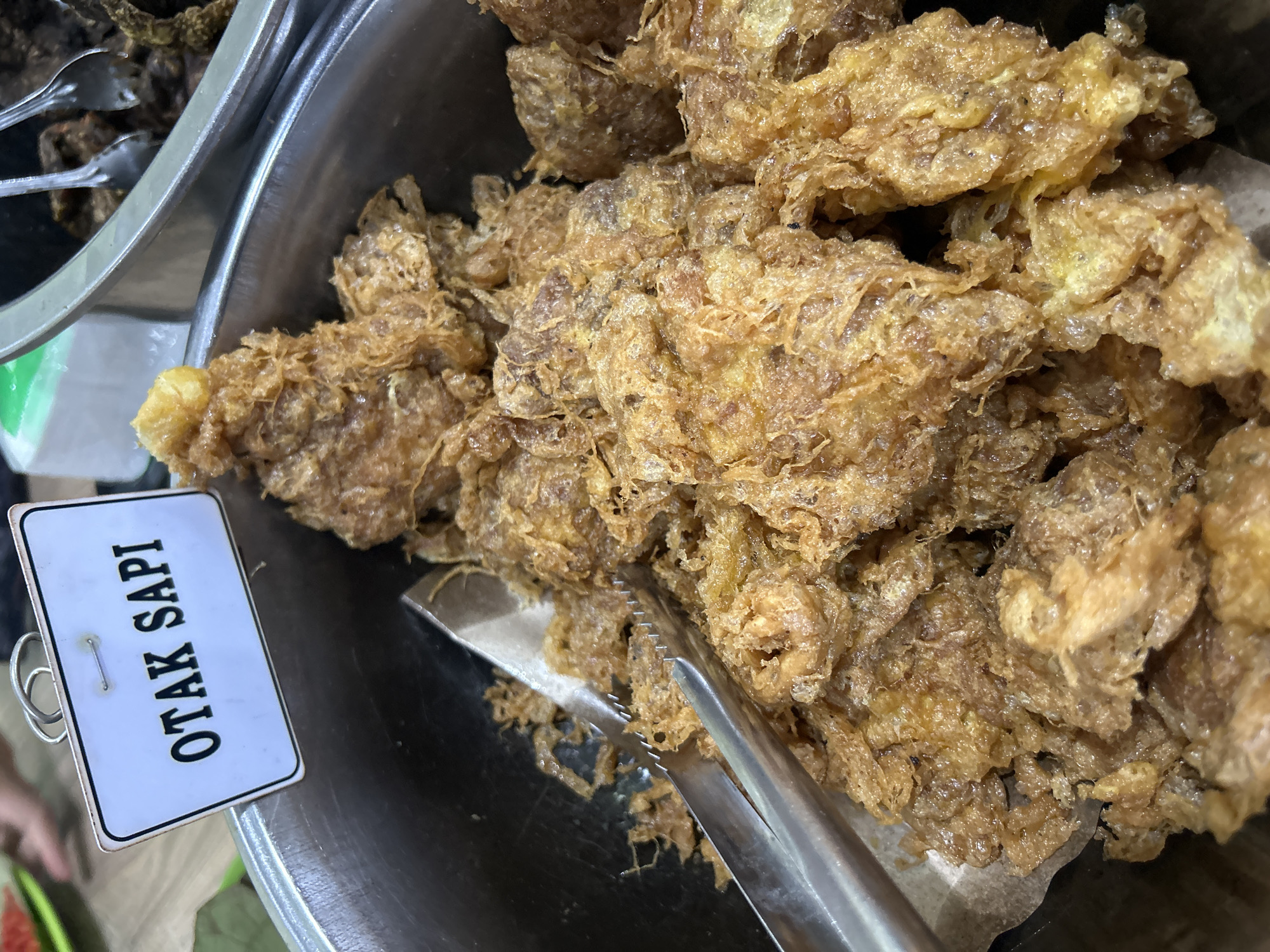 The image is a close-up, top-down view of a metal bowl filled with fried food, labeled "OTAK SAPI." The food appears to be pieces of meat or offal, battered and deep-fried, resulting in a crispy, golden-brown exterior. Tongs are positioned in the bowl, resting on the food. In the background, other food items and serving utensils are visible, suggesting a market stall or a food service establishment. The sign, "OTAK SAPI," is in black lettering on a white rectangular card with a decorative border, attached to the bowl with a wire loop. The overall impression is of a prepared dish ready for serving. The presence of the sign "OTAK SAPI" (Indonesian for "cow's brain") indicates the dish is likely sold in Cirebon, Indonesia, a city known for its diverse culinary scene.