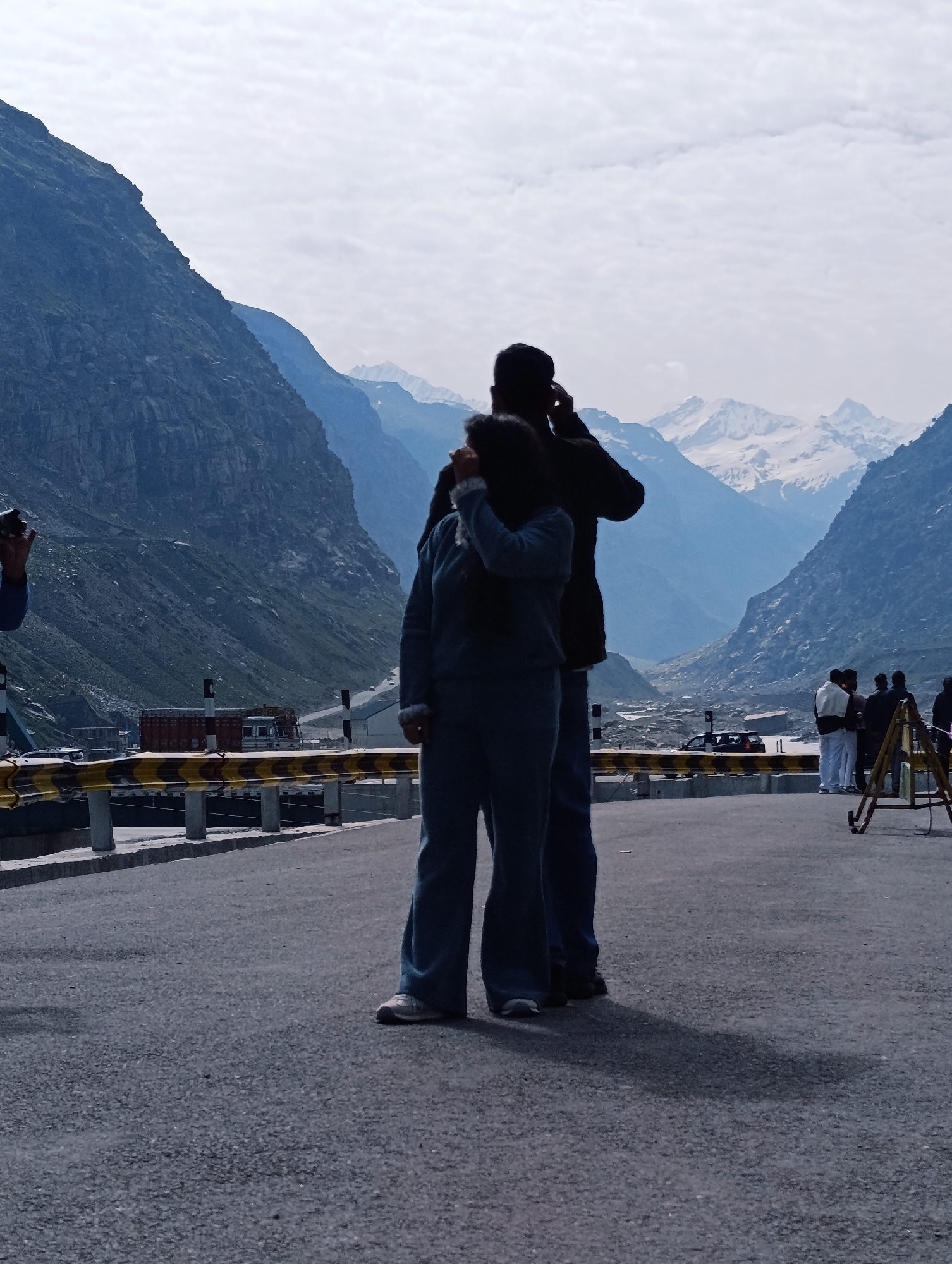 Two individuals, silhouetted against a backdrop of majestic, snow-capped mountains and a partly cloudy sky, stand on a paved road. The person in the foreground, wearing a blue tracksuit, is captured in profile with their hand covering their mouth, appearing to be speaking or reacting to something. Beside them, a second individual, also in silhouette, has their arm raised to their ear, as if on a phone call or listening intently. The mountainous terrain features steep, rocky slopes in the mid-ground, transitioning to distant, hazy peaks under a bright sky. In the background, a road winds through the valley, with a few vehicles visible, including trucks and a black SUV. Several other people are gathered further down the road, near a yellow and black striped barrier. The scene suggests a roadside stop in a mountainous region, possibly a scenic overlook or a point of interest along a travel route in India.