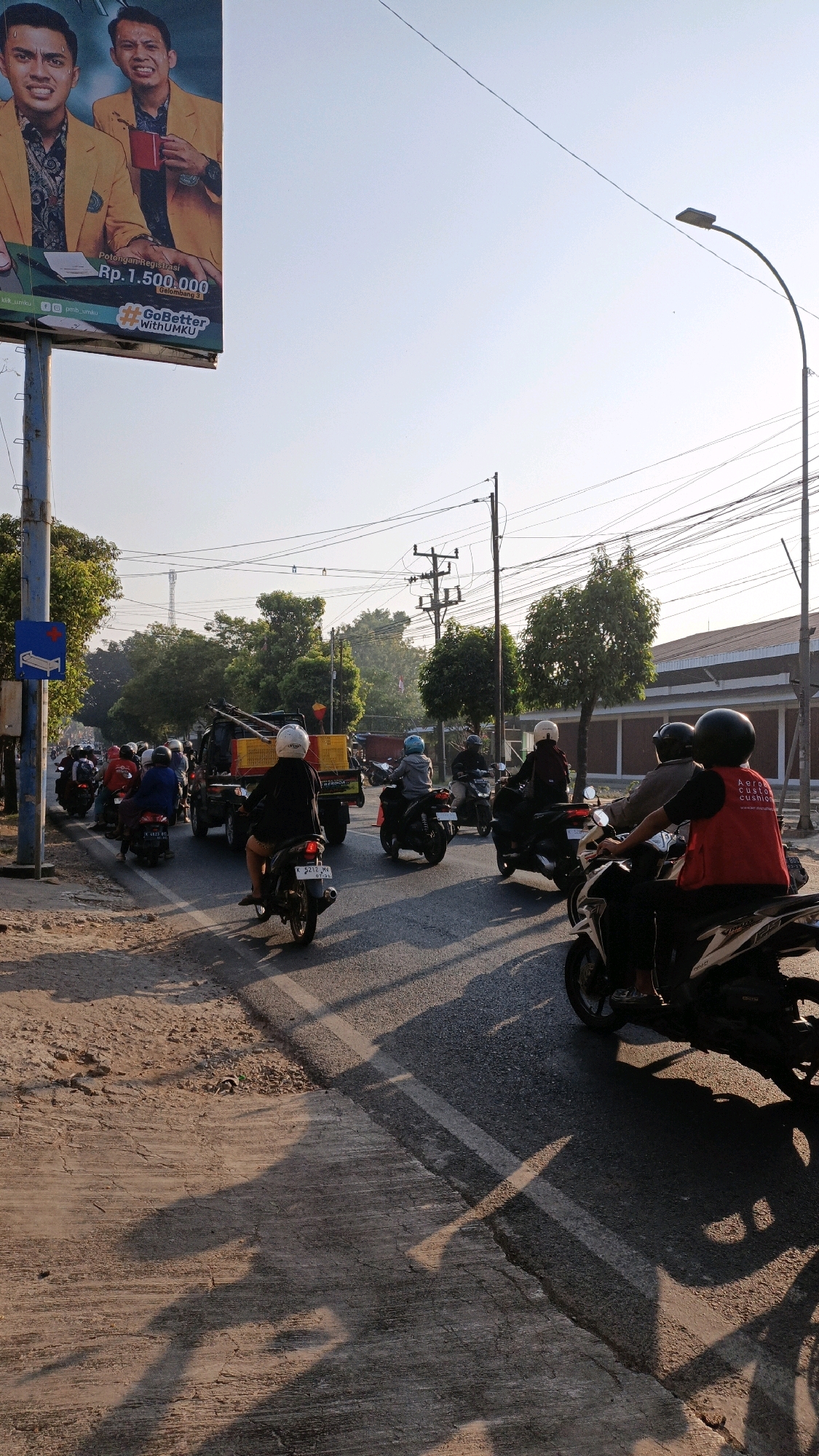 A street scene in Kuwasen, Indonesia, captures a moment of moderate traffic congestion during daylight hours. The foreground displays a textured concrete and dirt roadside, cast with long shadows from the setting or rising sun. The primary focus is a busy asphalt road where numerous motorcycles are navigating.
Several individuals are seen riding motorcycles. A motorcyclist wearing a red vest with white lettering, identified as "Aero Custom Cushion," is prominent in the right-hand side of the frame. Other riders are visible, many wearing helmets in various colors like white, blue, and black. Some motorcycles are stationary or moving slowly, indicating a traffic jam.
A yellow flatbed truck with a metal frame is stopped in the middle of the road, carrying a yellow container. A traffic sign depicting a blue square with a white medical cross symbol is positioned on a pole to the left of the road. In the background, trees line the street, and power lines crisscross overhead, supported by utility poles. A large billboard is partially visible on the far left, featuring two men in yellow jackets and text that includes "Rp.1.500.000" and "GoBetter WithUMKU." The sky is clear and bright, suggesting fair weather. The overall impression is one of an active urban environment with typical daily traffic conditions.