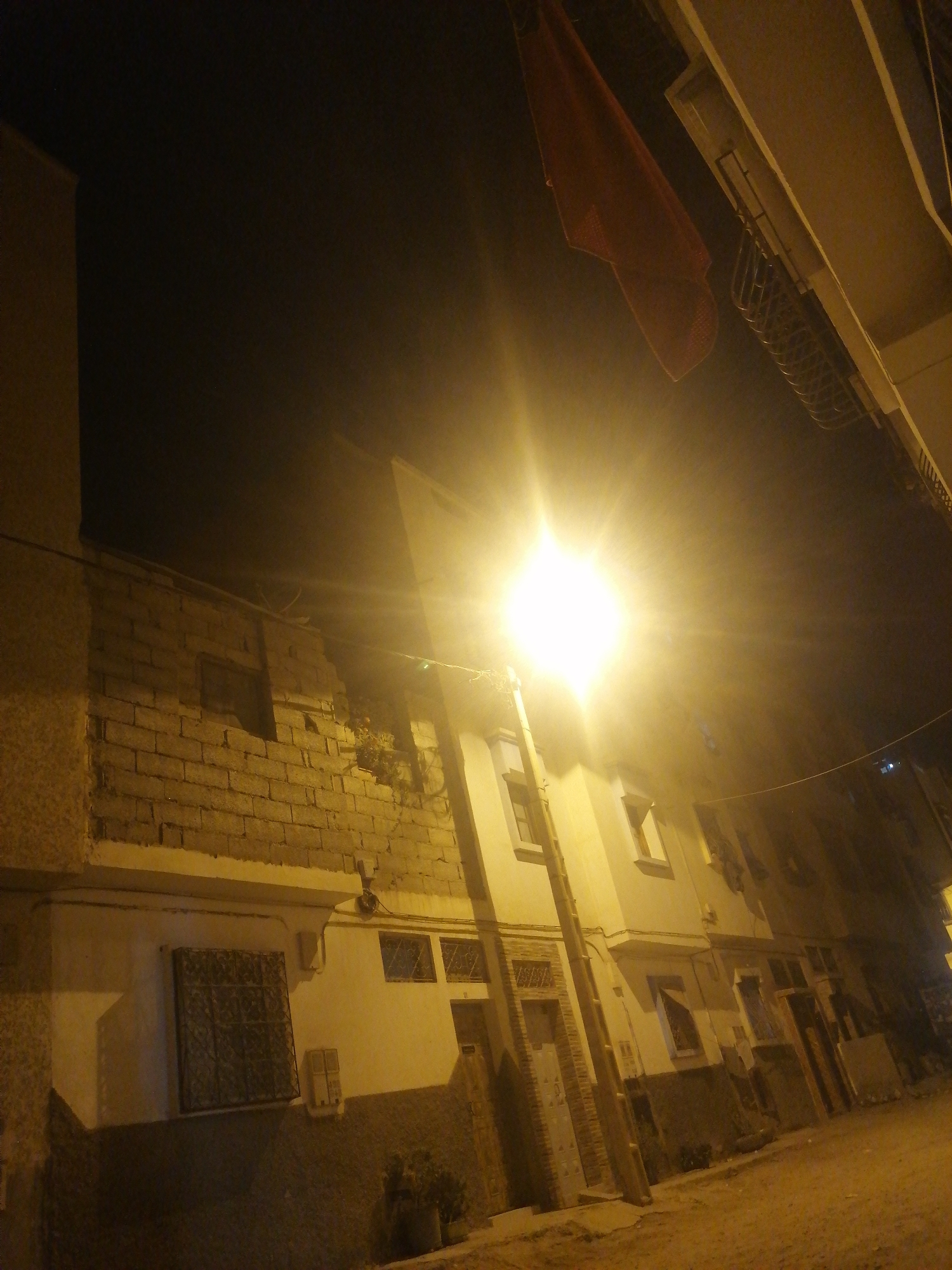 The image depicts an outdoor scene at night, possibly in a city. The foreground shows a street or alleyway, with the ground appearing to be dirt or gravel. To the left, there is a textured wall, possibly made of concrete. Further back, a building is visible, constructed with brickwork. The building has several windows, some of which are barred. A bright light source, possibly a streetlamp, is prominent in the upper center of the image, casting a strong glare. In the upper left quadrant of the image, a red fabric object, possibly a flag or banner, is suspended from a wire and appears to be blowing in the wind. The overall lighting is dim, with the bright lamp creating high contrast and washed-out areas. The scene appears to be deserted.