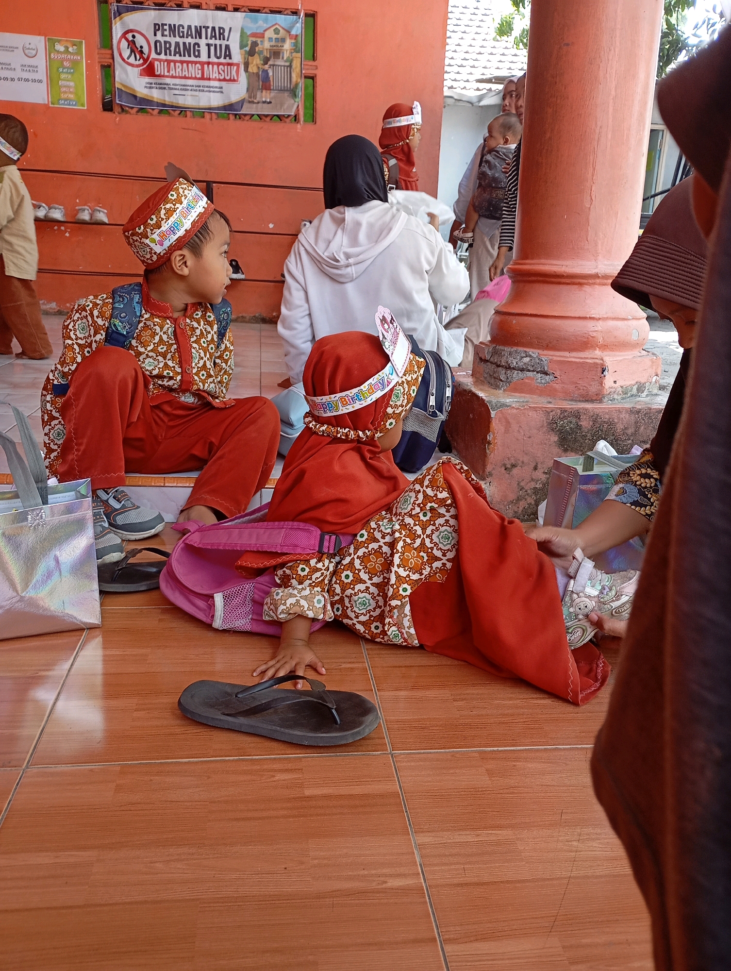 In the city of Ketegan, Indonesia, a group of children and adults are gathered outside an orange-colored building with a tiled floor. Two young children, a boy and a girl, are seated on the floor, both wearing traditional Indonesian clothing. The boy, to the left, is dressed in a red and white patterned shirt and pants, with a matching hat adorned with "Happy Birthday" text. He has a blue backpack on. The girl, to the right, wears a red outfit and a matching headpiece. She is lying on her side, seemingly interacting with a pair of light-colored sneakers. A pink backpack is positioned behind her. In the background, several adults are visible, some holding infants, and a sign with Indonesian text is displayed on the wall. The environment appears to be an outdoor space adjacent to a building, possibly a school or community center.