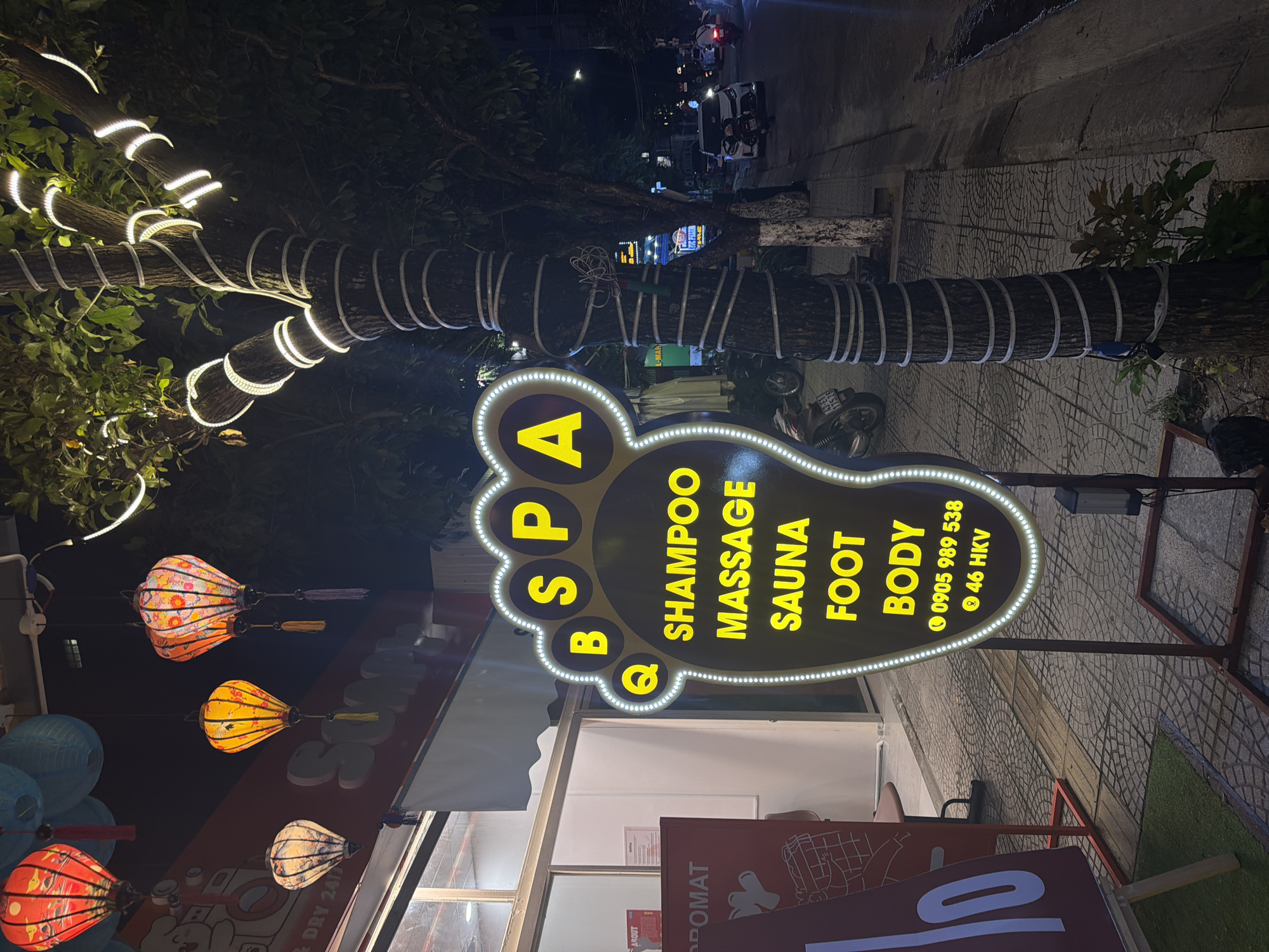 A nighttime shot of a street in Đà Nẵng, Vietnam. The focus is on a sign for a spa called "QB SPA" shaped like a foot, listing services such as "SHAMPOO, MASSAGE, SAUNA, FOOT, BODY," along with a phone number (0905 989 538) and address (46 HKV). Above the sign, a tree's trunk and branches are illuminated with white string lights wrapped around them. Several colorful lanterns hang in front of a building, possibly the spa itself. There are motorbikes and cars visible further down the street, indicating moderate traffic. The ground is paved, and the sign is on a stand with metal support. A sign that reads 'PROMAT' is visible at the bottom.