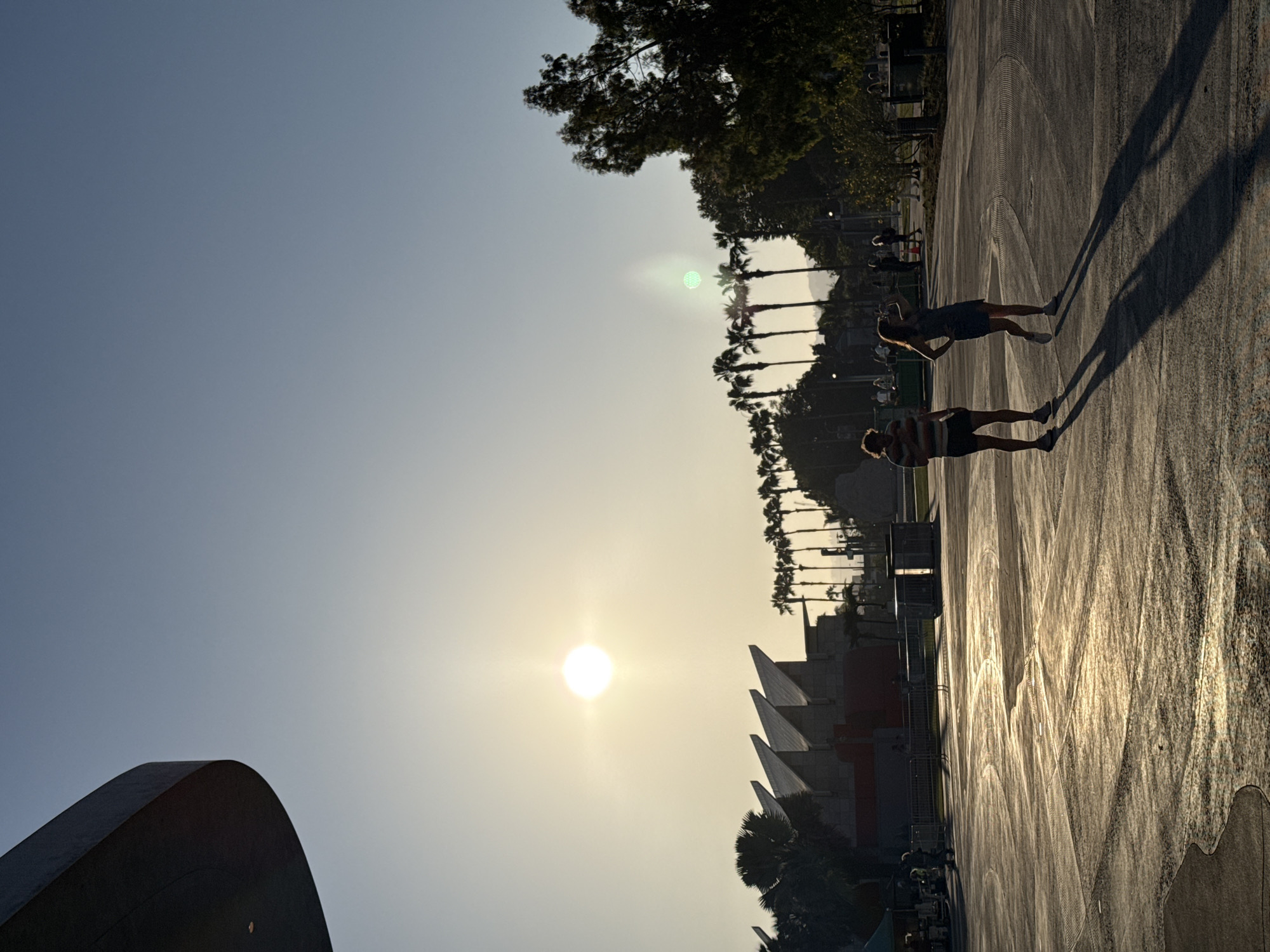 This image captures an outdoor scene in Los Angeles, USA, likely during the late afternoon or early evening, given the low angle of the sun and the long shadows. The sun is a prominent feature, positioned in the upper left quadrant of the sky, casting a bright, diffused light across the scene.
In the foreground, a paved area with circular markings is visible, with two individuals silhouetted against the bright ground. One is a male wearing a striped t-shirt and shorts, and the other is a female in a dress. They are standing and facing away from the camera, with their long shadows stretching out towards the viewer.
In the mid-ground, a row of palm trees stretches horizontally across the image. Behind the palm trees, there is a distinctive building with a pointed, angular roofline, illuminated by the sun. Further in the background, more trees and distant hills are visible.
The overall environment appears to be an open public space, possibly a park, plaza, or an area adjacent to a cultural institution or entertainment venue, characterized by its modern architecture and natural elements like palm trees. The presence of a few other silhouetted figures and what appear to be temporary structures or stalls on the far left suggest some activity or event is taking place.