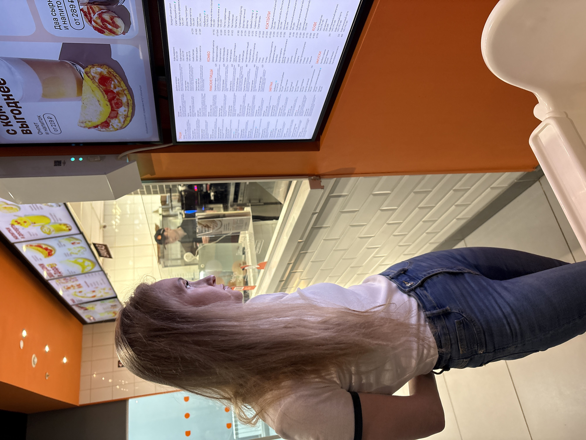 The scene depicts the interior of a modern fast-casual restaurant in Izhevsk, Russia. In the foreground, a blonde female, estimated to be between 20-30 years old, stands in profile, facing right. She has long, straight hair and is wearing a white short-sleeved t-shirt with a dark band on the left sleeve and blue denim jeans. Her posture indicates she is intently viewing digital menu boards.
To the right, two large, vertically oriented digital display screens are mounted on an orange wall section. The upper screen shows images of food and drink combinations, including an omelet with a latte-style beverage priced "от 219 Р" (from 219 Rubles). The lower screen displays a comprehensive text menu in Cyrillic, categorized into "Пиццы" (Pizzas), "Комбо" (Combos), "Завтрак" (Breakfast), "Закуски" (Snacks), "Коктейли" (Cocktails), and "Кофе" (Coffee). A white rectangular device with two illuminated blue lights is affixed to the wall next to the upper menu board. Partially visible in the lower right is the back of a white high chair.
In the background, behind a white counter and through a transparent barrier, a male employee in a dark uniform and cap is visible in what appears to be a kitchen or food preparation area, engaged in tasks. The ceiling features orange panels with recessed spotlights. A white subway-tile-style wall section is present in the mid-ground, below the counter. The overall environment is brightly lit and clean, consistent with a commercial food service establishment.